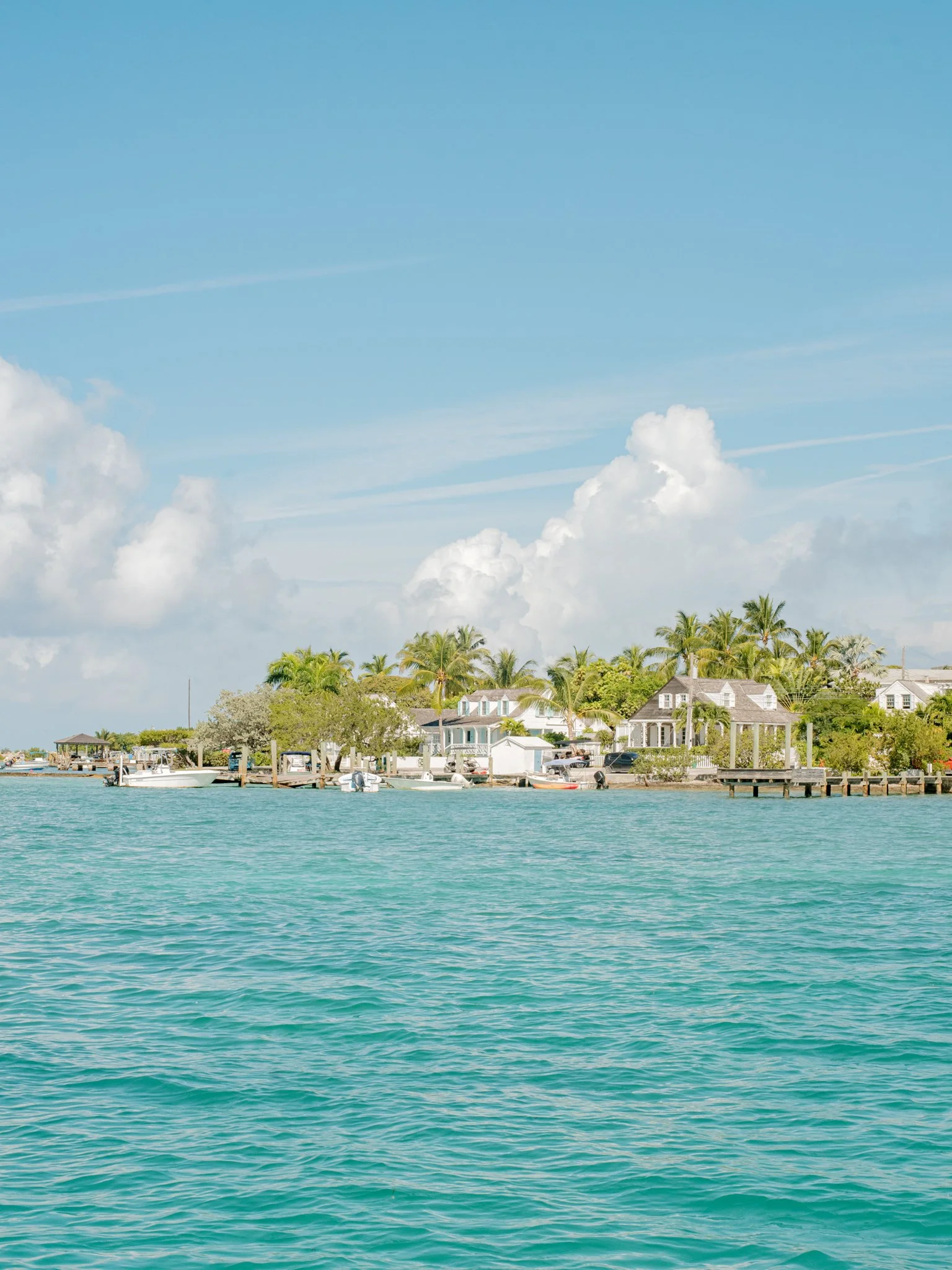 A waterfront scene with houses, palm trees, boats, and clear blue water under a partly cloudy sky.