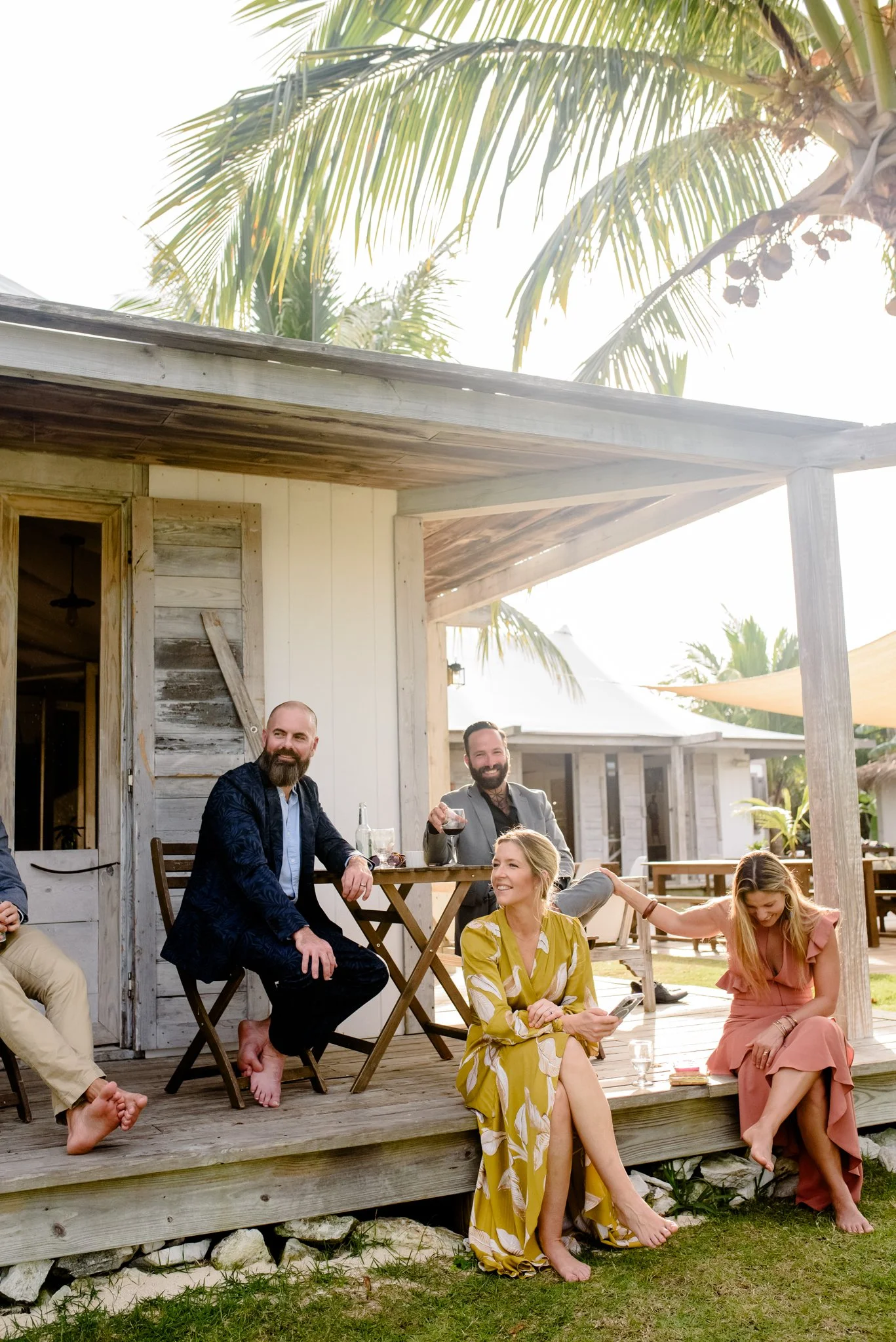 Group of friends enjoying drinks and conversation on a wooden porch of a beach house surrounded by palm trees.