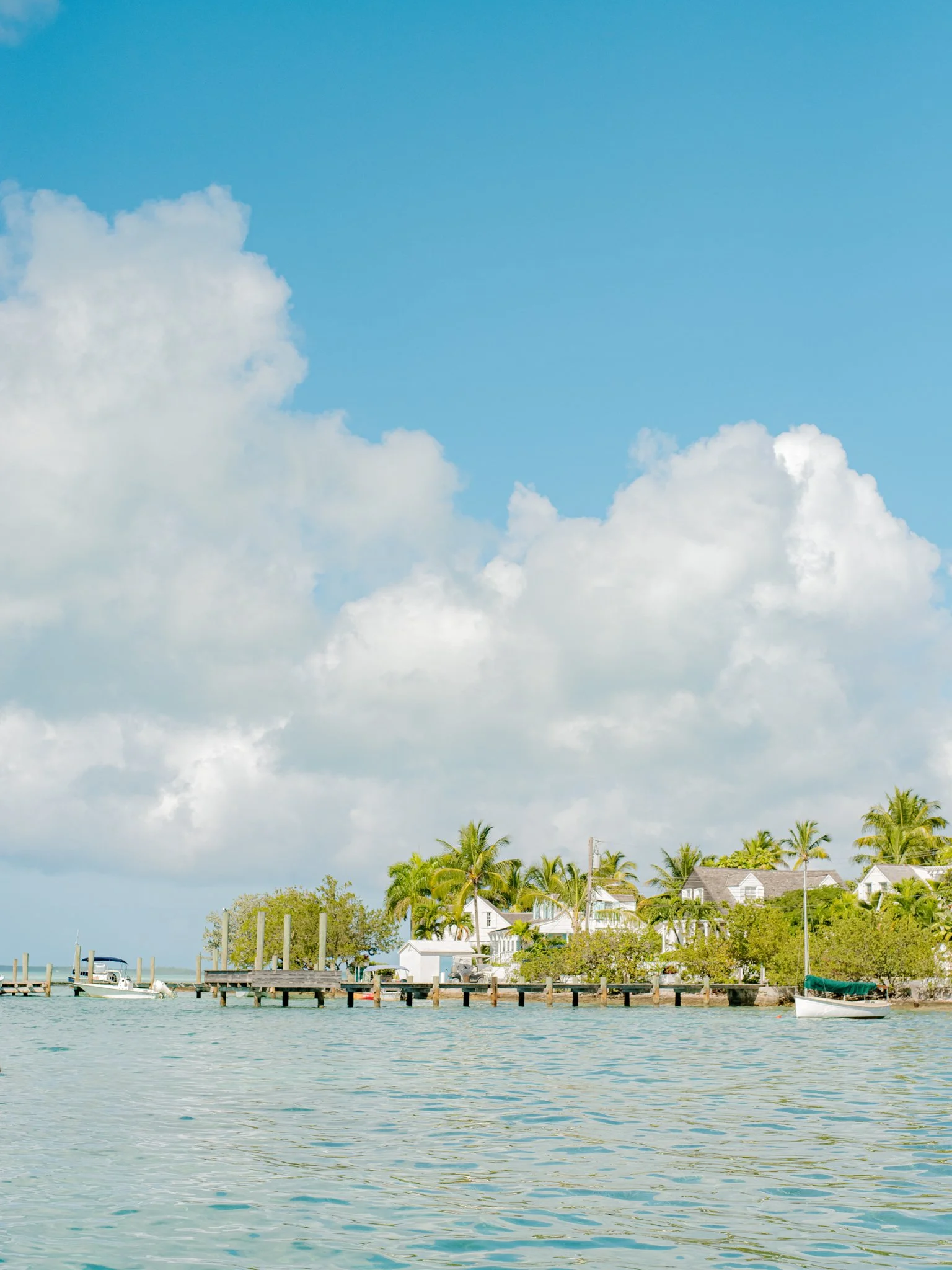 A scenic view of a waterfront with small boats, a dock, lush green trees, and houses with palm trees under a partly cloudy sky at The Other Side Hotel during their destination wedding in Eleuthera, Bahamas.