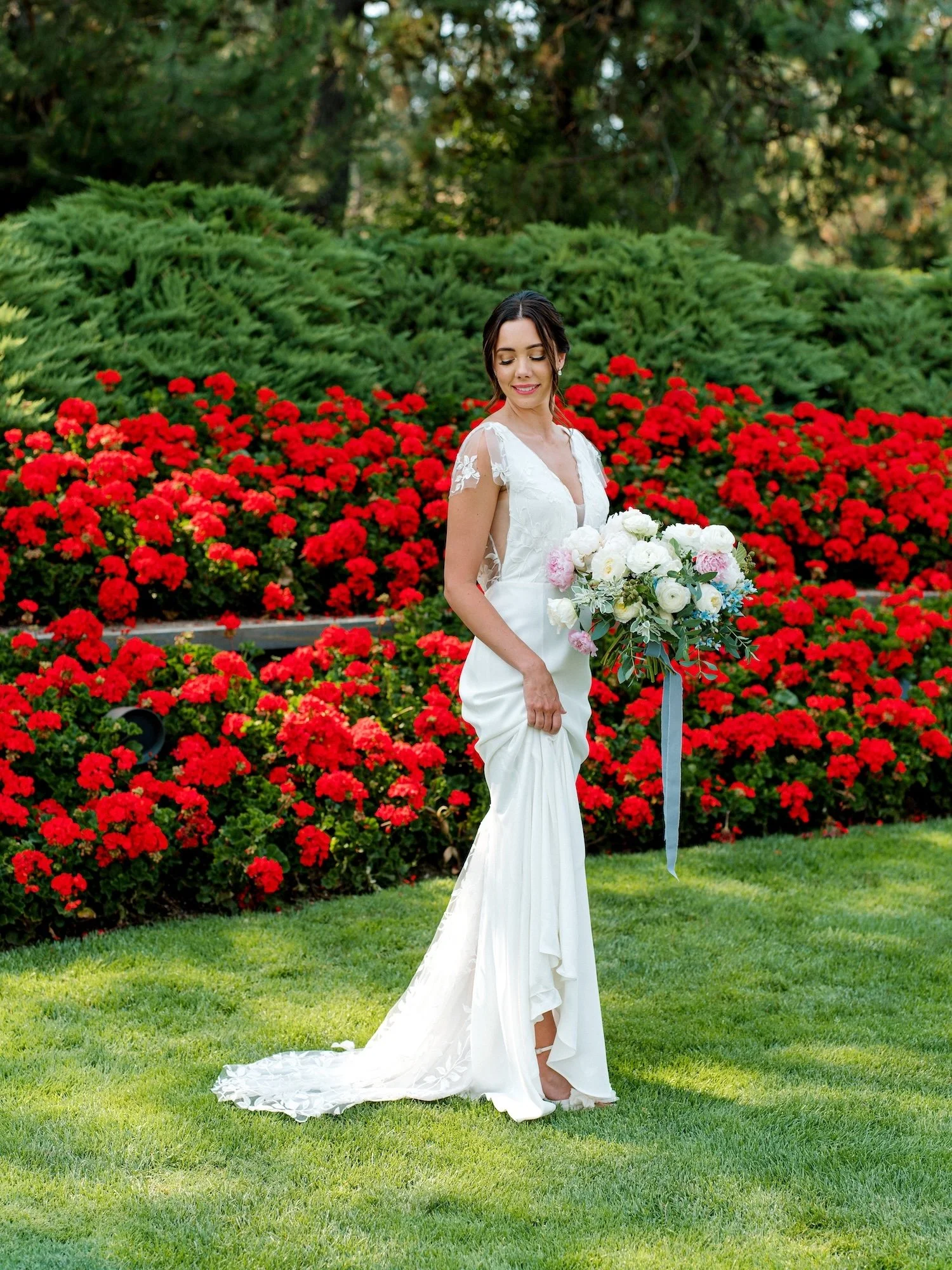 Bride in a white wedding gown holding a bouquet of white, pink, and blue flowers, standing outdoors in front of a colorful garden with red flowers and green bushes.