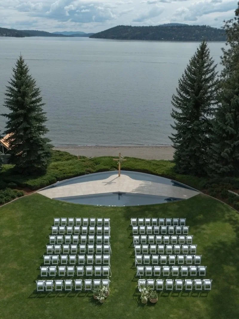 Outdoor wedding setup with white chairs facing an altar with a cross, overlooking a lake with pine trees and hills in the background.