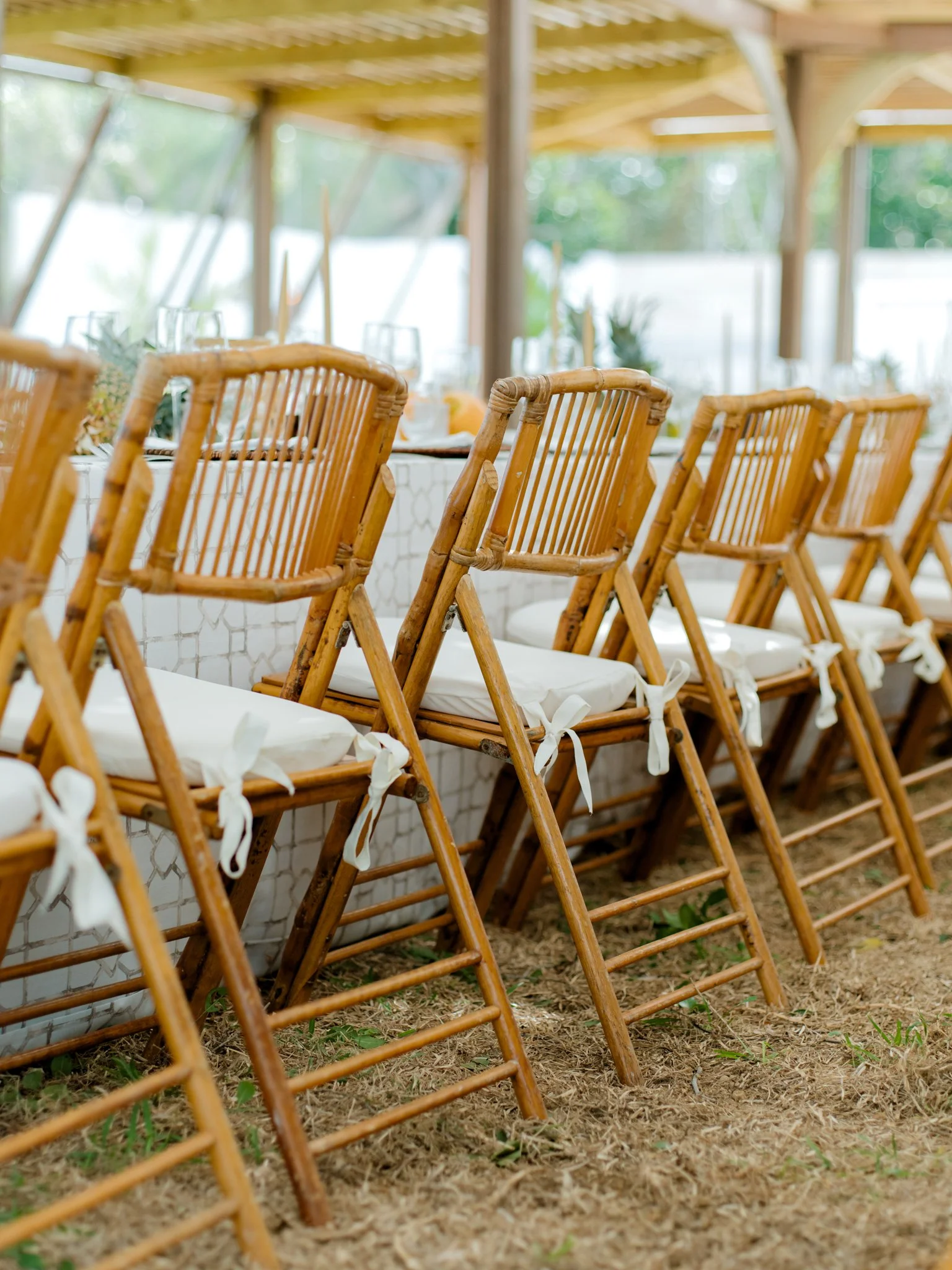 Row of bamboo chairs with white cushions tied to a long dining table at an outdoor event space.