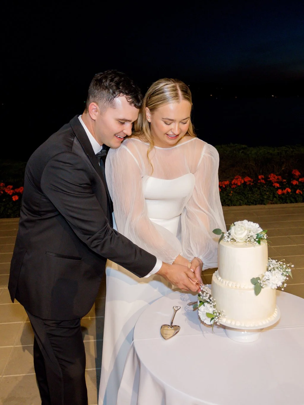 Happy couple in wedding attire cutting a white wedding cake decorated with white flowers and greenery outdoors at night.