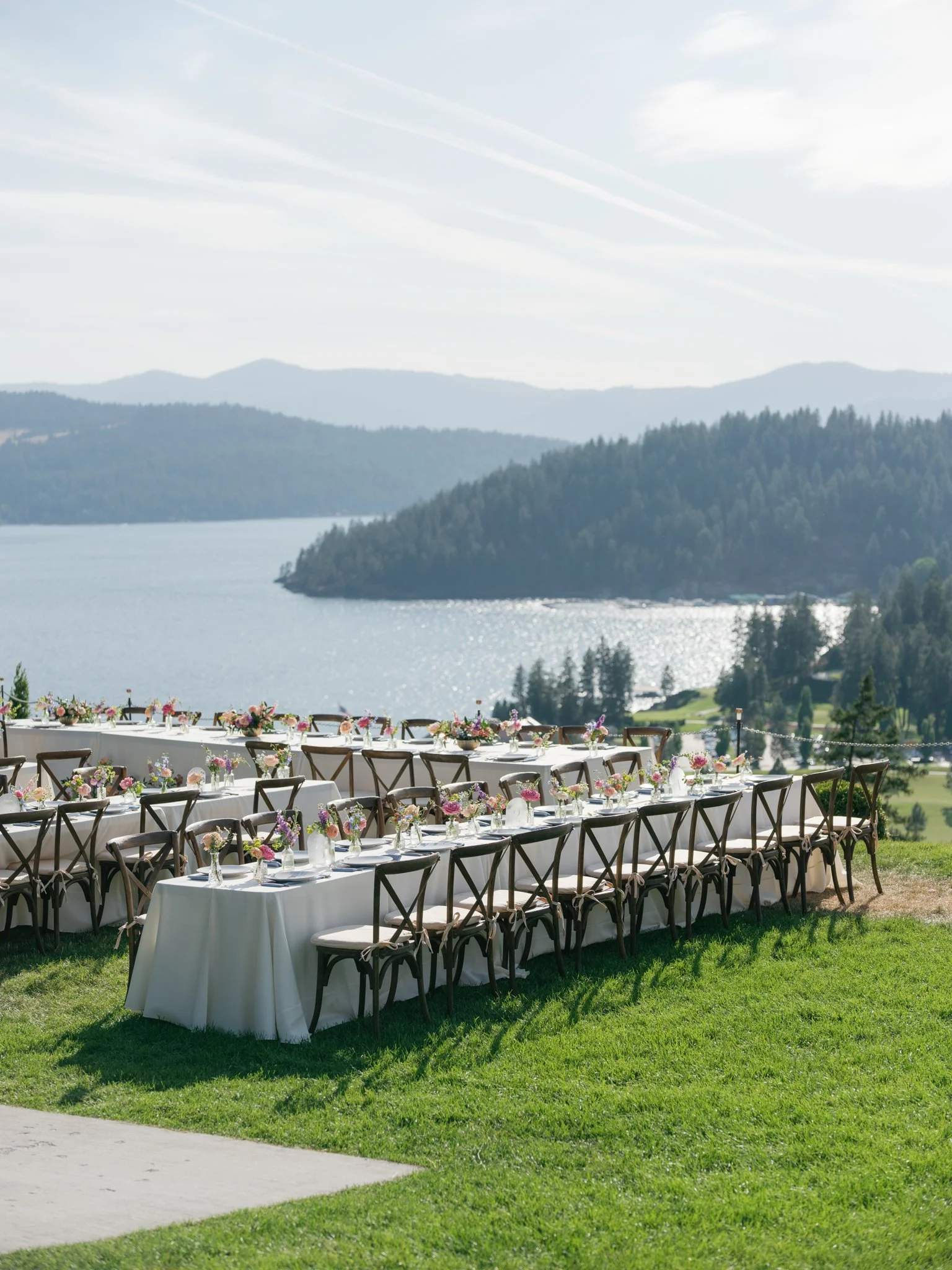 Long banquet table set for outdoor event with white tablecloths, floral centerpieces, and chairs, overlooking a lake surrounded by trees and mountains under a partly cloudy sky at Elk Point wedding in Coeur d’Alene.