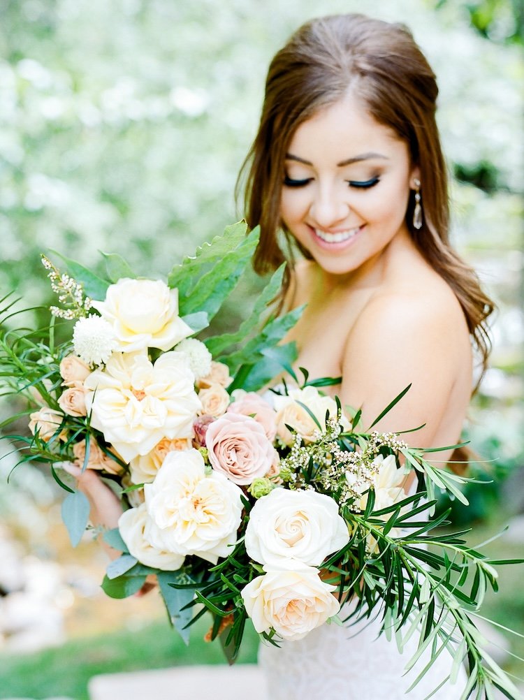A woman on her wedding day holding a large bouquet of flowers, including roses and greenery, outdoors with a blurred green background.