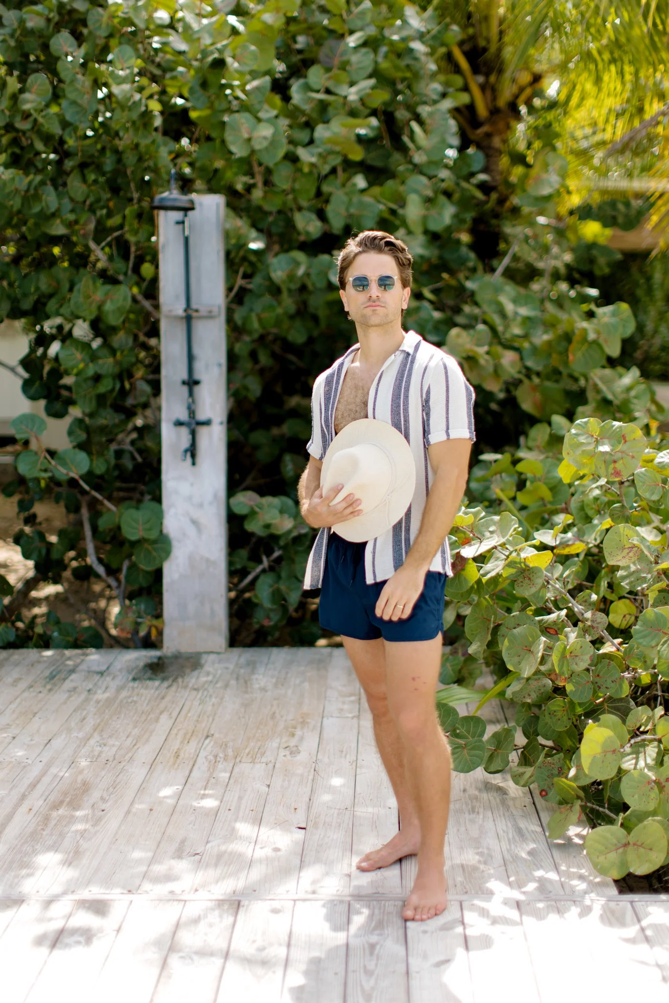 A young man with sunglasses, wearing a striped shirt, navy shorts, and holding a wide-brimmed hat, standing barefoot on a wooden deck with lush green foliage in the background. The Other Side Hotel in Eleuthera.