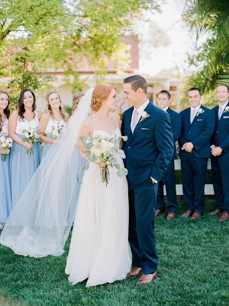 A bride and groom face each other smiling during their wedding ceremony outdoors, surrounded by bridesmaids and groomsmen, with green trees and a building in the background.