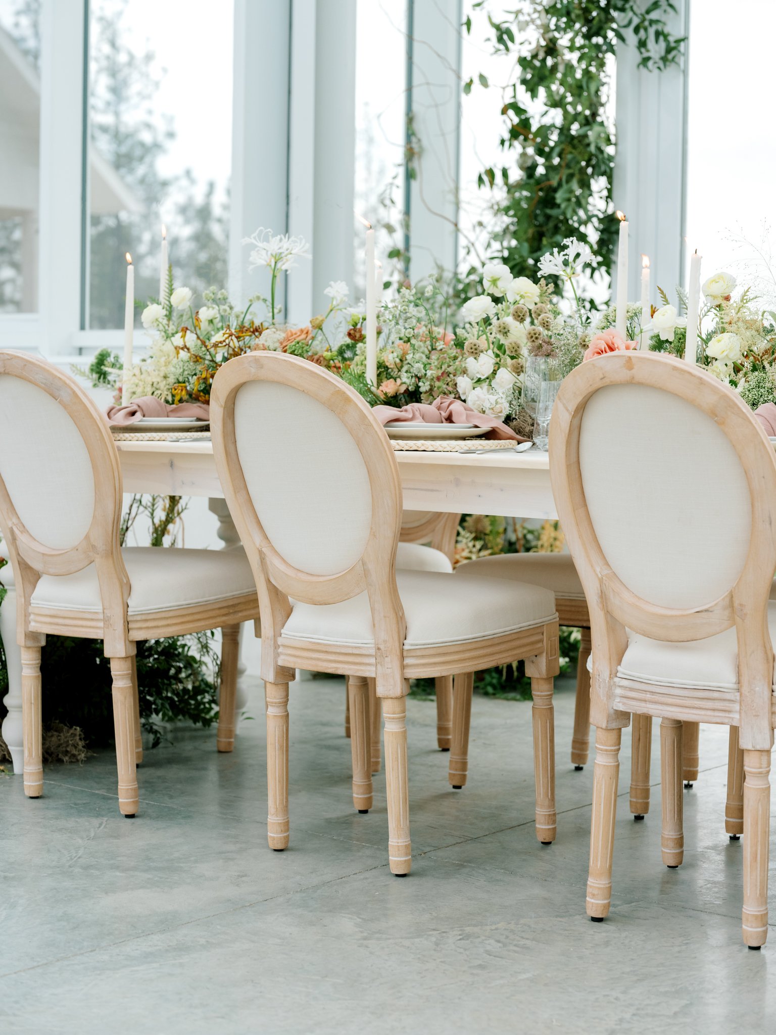 Elegant dining table set with floral centerpiece, candles, and cream-colored chairs in a glass-enclosed space with greenery outside at Greenhouse on Harvard near Spokane, Washington