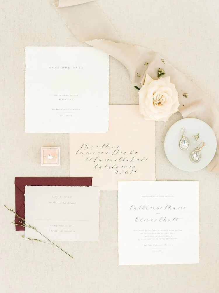 Wedding invitation suite with white and cream cards, a flower, a silk ribbon, a ring in a pink box, and a pair of teardrop earrings.