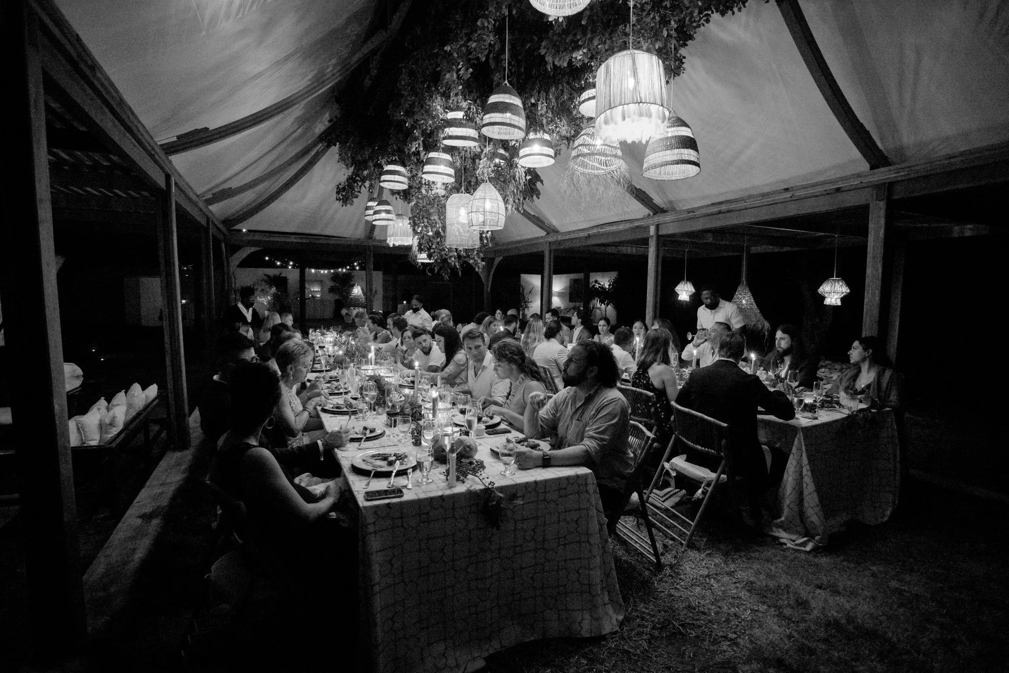 A black-and-white photo of a large outdoor dining gathering inside a tent with hanging lamps and decorative foliage at The Other Side Hotel during their destination wedding in Eleuthera, Bahamas.