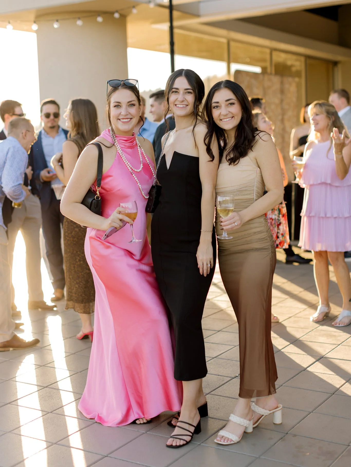 Three women dressed in elegant evening gowns smiling and holding wine glasses at an outdoor gathering during sunset with other people in the background.