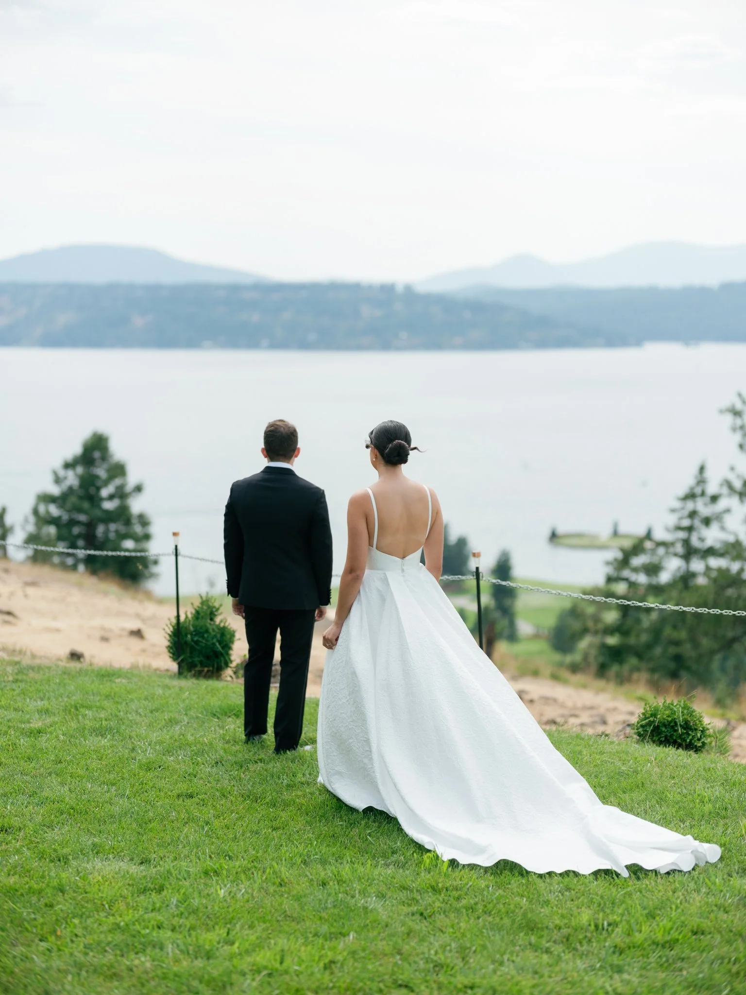 A bride and groom standing outdoors on a grassy hill, facing a lake with mountains in the background, during their wedding ceremony at Elk Point mountaintop wedding.