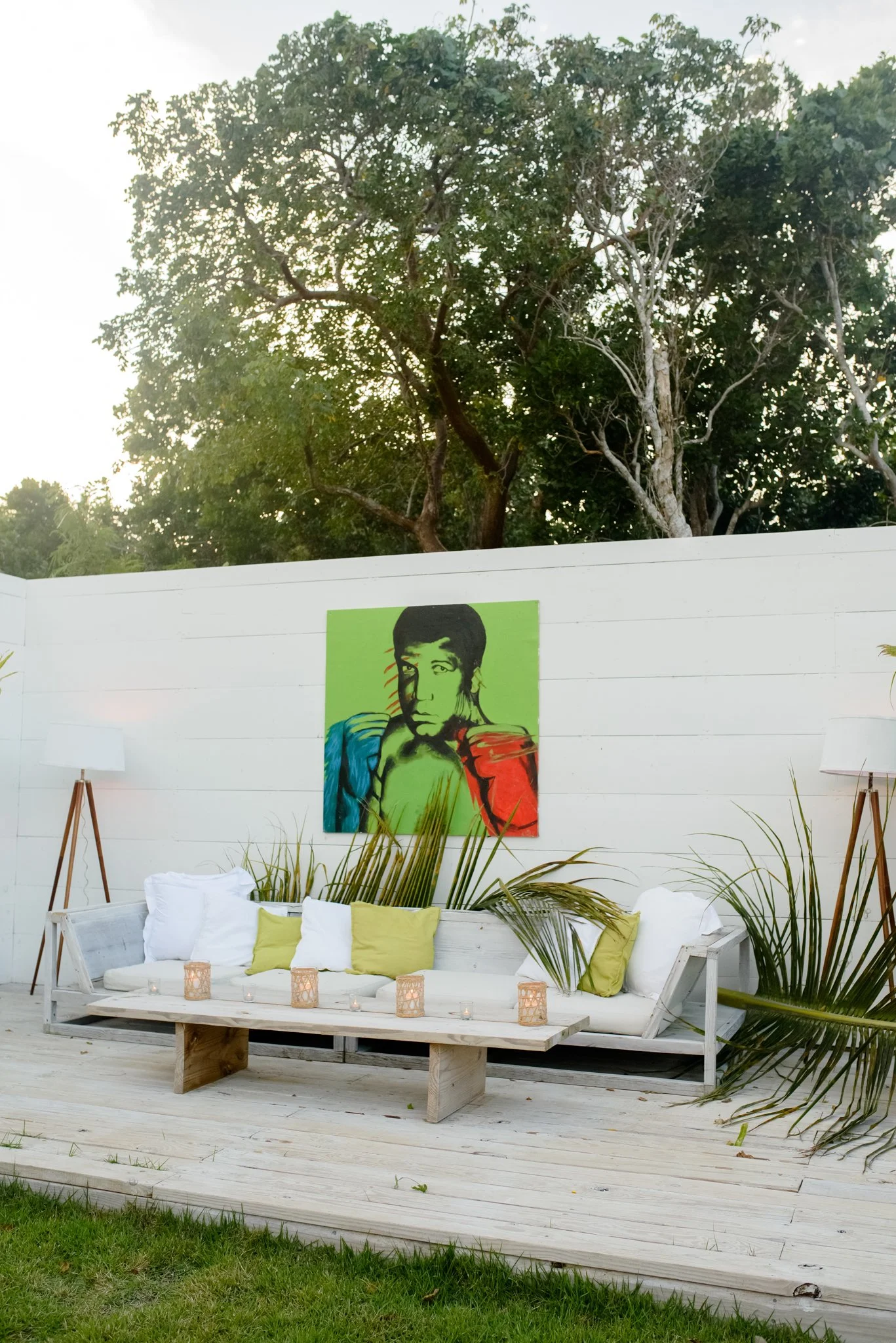 Outdoor patio with a white wooden couch with white and light green pillows, a rustic wooden coffee table with candles, two tall floor lamps with white shades, and a colorful pop art painting of a woman on the white fence behind the couch. Green plants and trees are visible in the background.