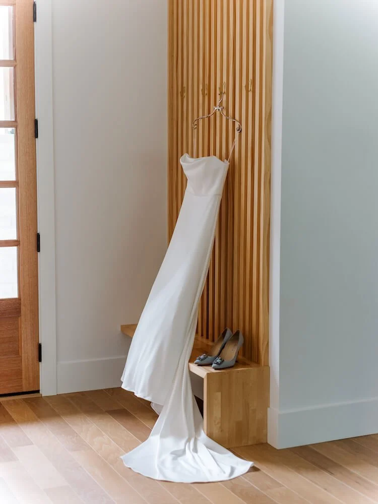 A white wedding dress hanging on a hook beside a pair of gray high heels on a wooden shelf, against a wooden slat wall in a room with hardwood flooring.