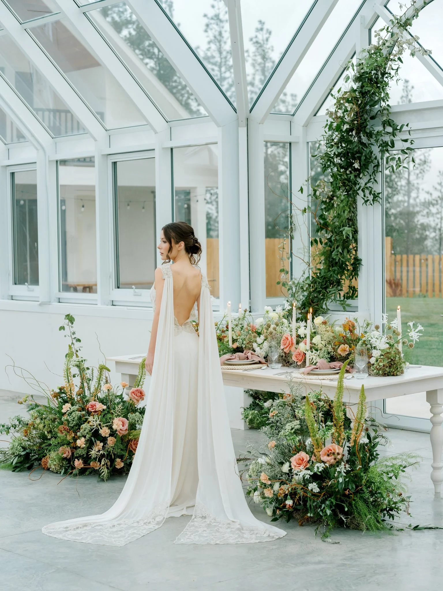 A bride in a backless white wedding gown with flowing train stands in a greenhouse surrounded by floral arrangements and candles on a white table at Greenhouse on Harvard near Spokane, Washington.