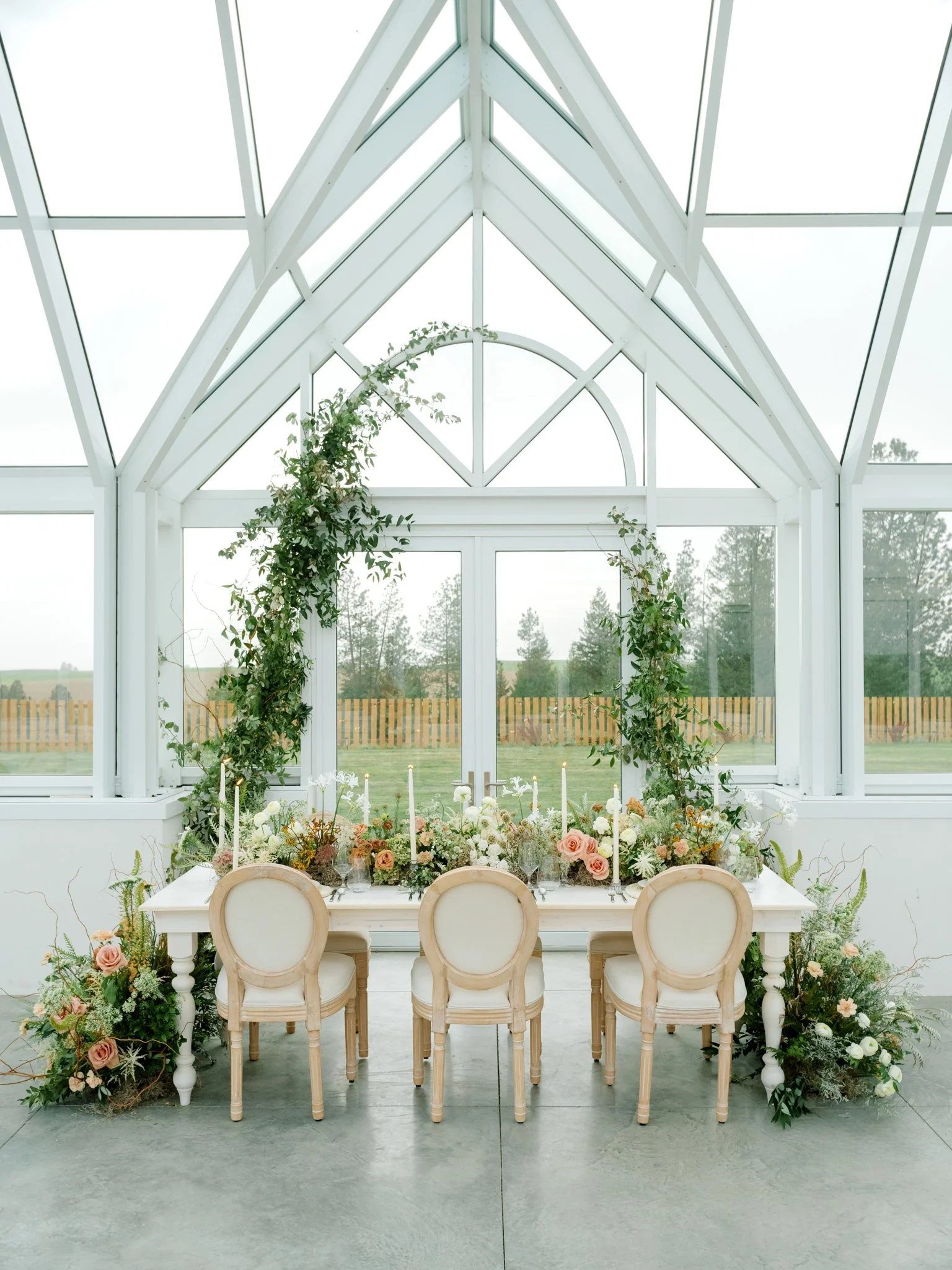 A wedding or event table setup inside a bright, glass conservatory with white framing, decorated with flowers and greenery, and surrounded by chairs at Greenhouse on Harvard near Spokane, Washington