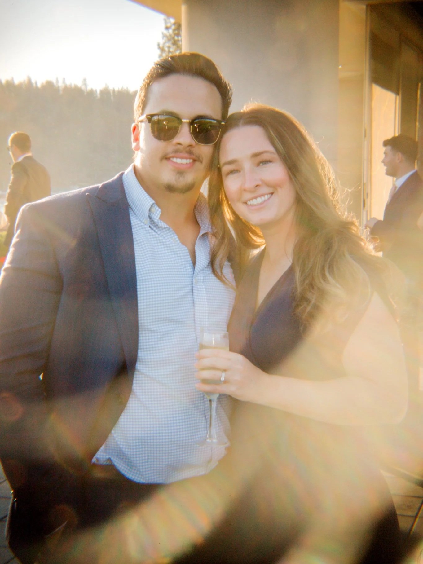 A man with sunglasses and a woman smiling, holding a champagne flute, standing outdoors at sunset during a social event.