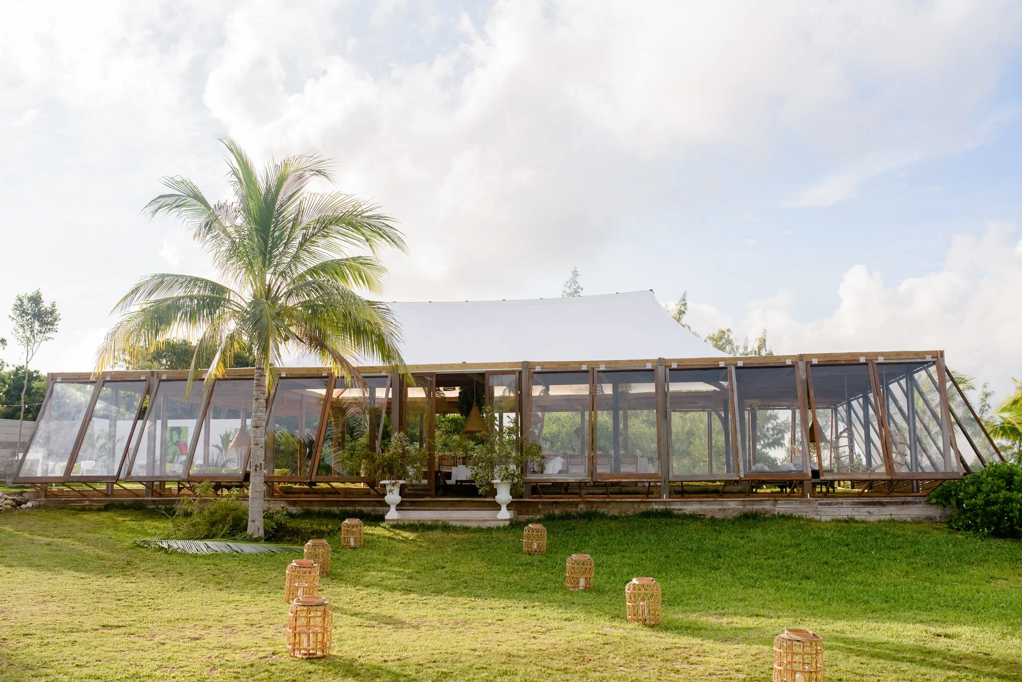 A tropical outdoor setting with a large white canopy tent on a wooden platform, surrounded by clear plastic walls and a grassy area. There is a tall palm tree to the left of the tent, and small decorative lanterns are placed on the grass in front of the tent.