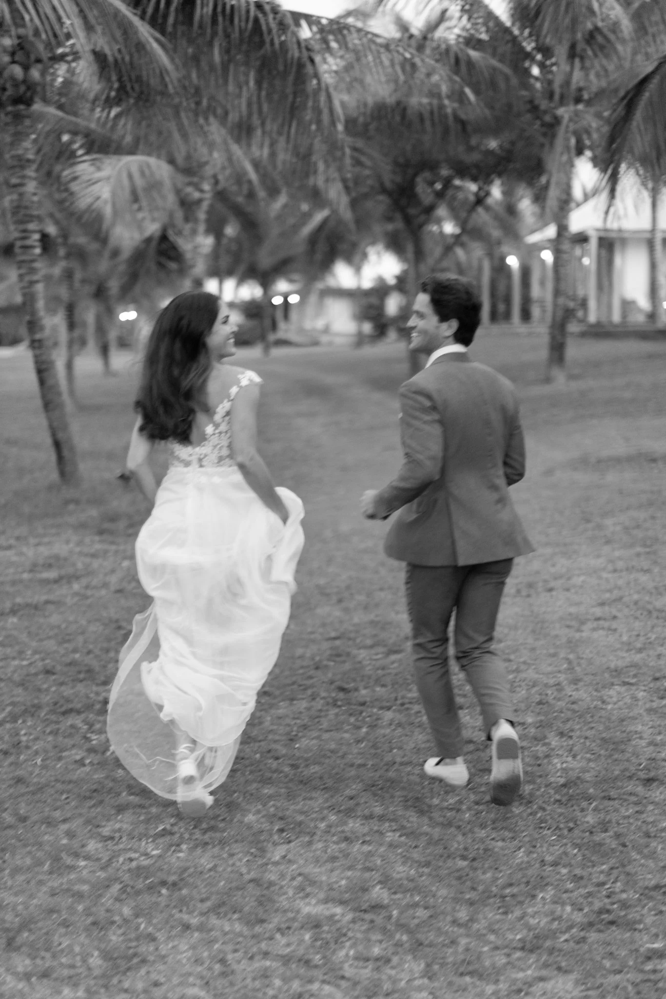 A bride and groom running and smiling in an outdoor park with palm trees, dressed in wedding attire, in black and white at The Other Side Hotel Eleuthera during a luxury Bahamas destination wedding.