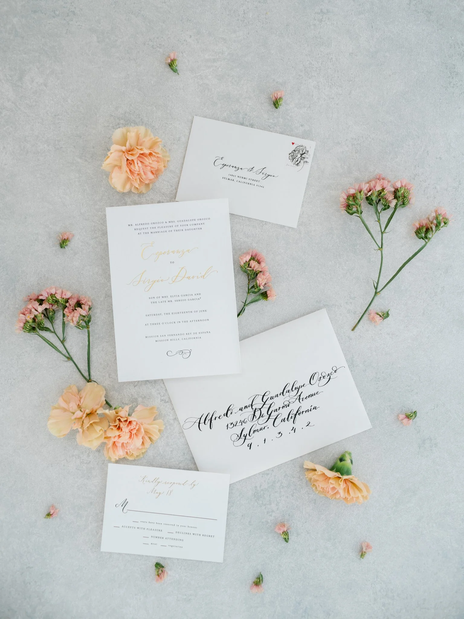 Wedding invitation stationery with floral decorations, including an invitation card, RSVP card, and envelopes, adorned with peach and pink carnations and small pink flowers on a light gray surface.