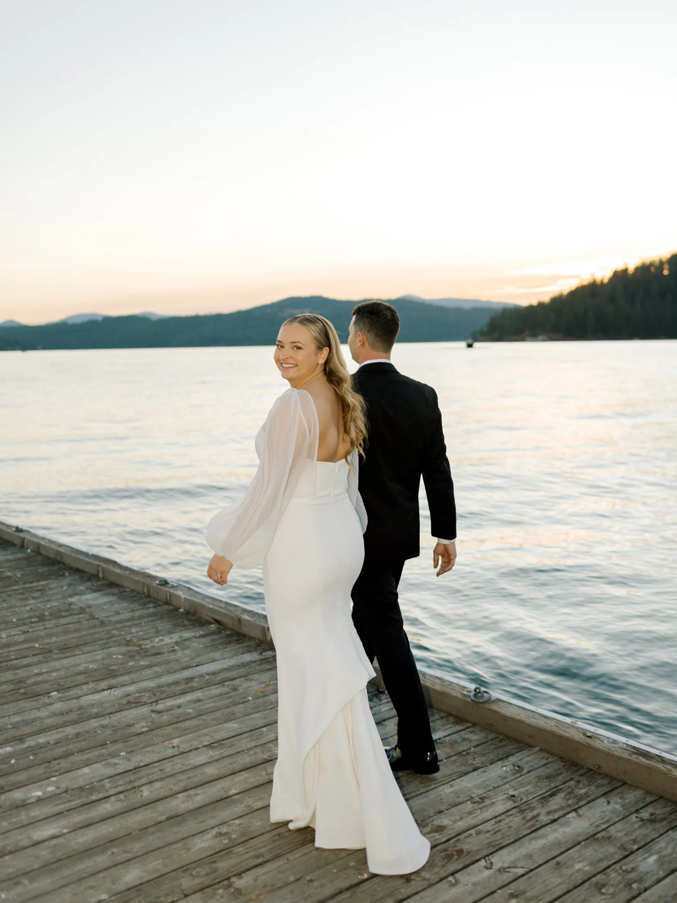 A bride and groom walking on a wooden dock by a calm lake at sunset, with a mountainous landscape in the background. The bride is smiling towards the camera, wearing a white wedding dress with long sleeves, and the groom is dressed in a black suit, walking away from the camera.