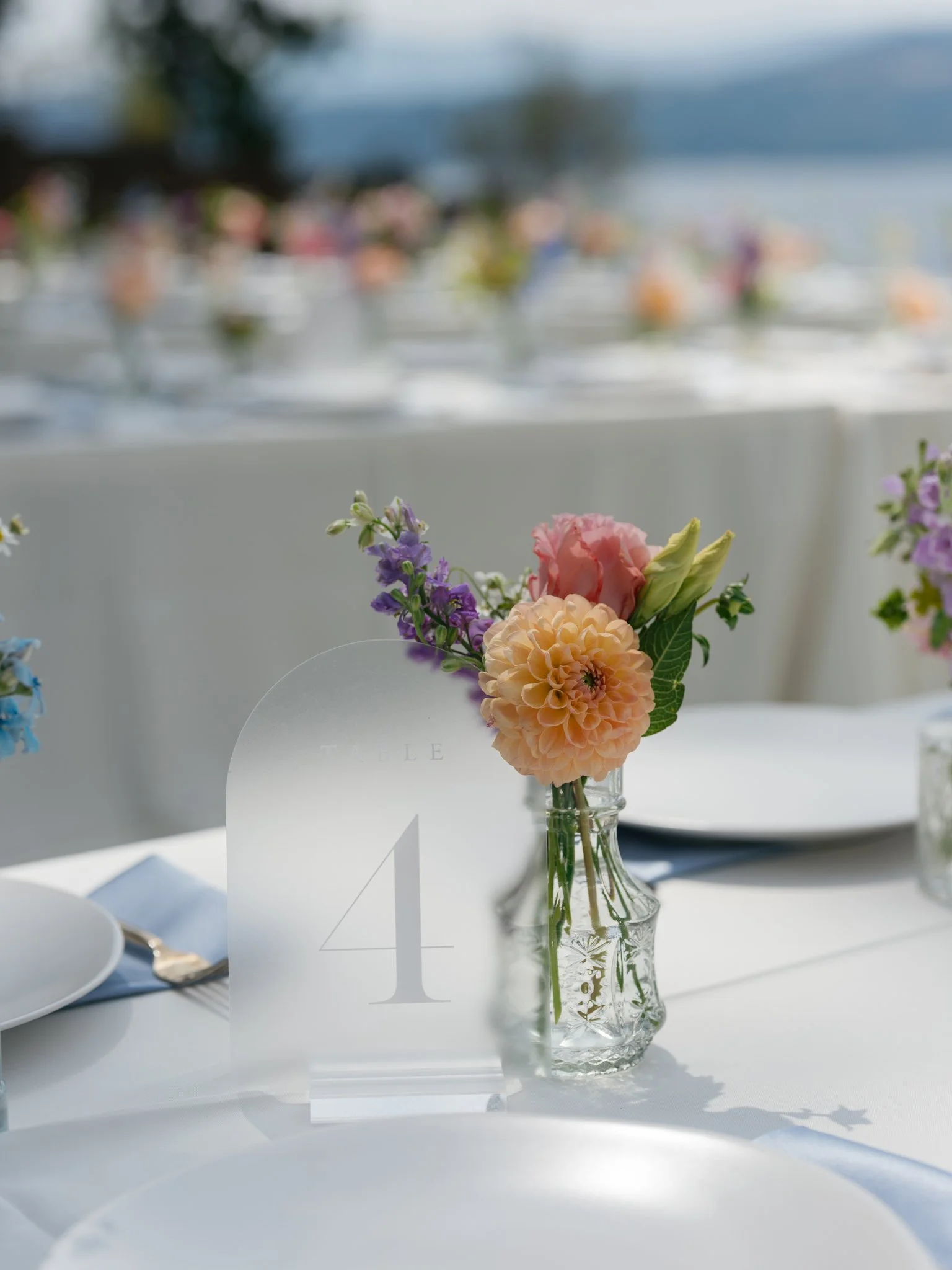 Elegant table setting with a small glass vase of pink, purple, and peach flowers, a white table card with the number 4, and white plates and silverware, outdoor event with blurred background of more tables on a lakeside at Elk Point wedding.