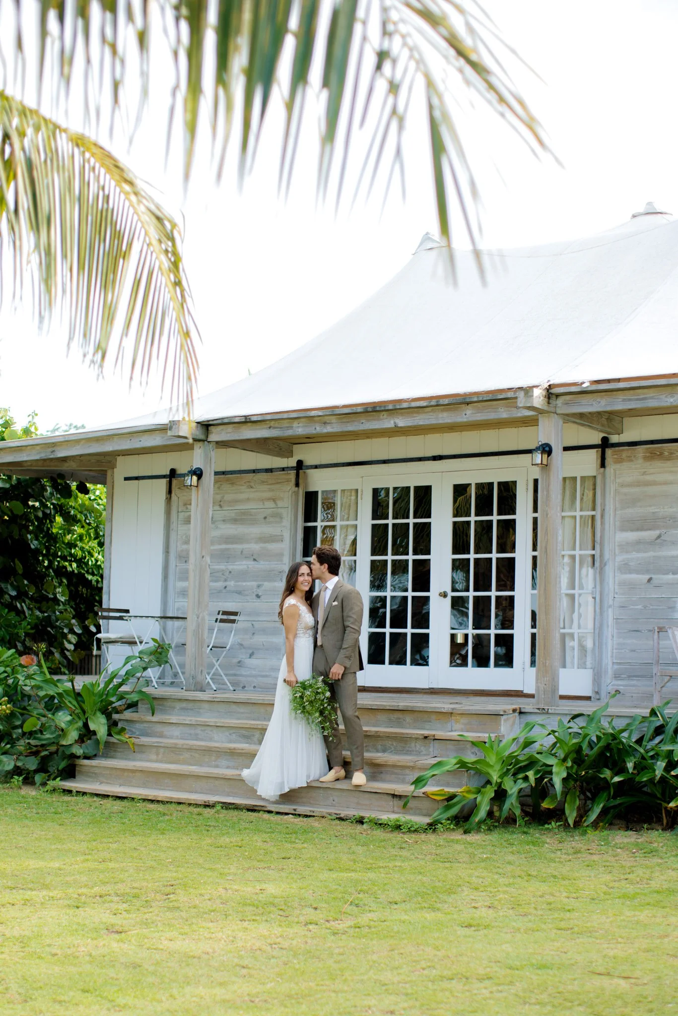 A bride and groom standing on the steps of a wooden house with glass doors, surrounded by greenery, with palm tree leaves hanging above at The Other Side Hotel Eleuthera during a luxury Bahamas destination wedding.
