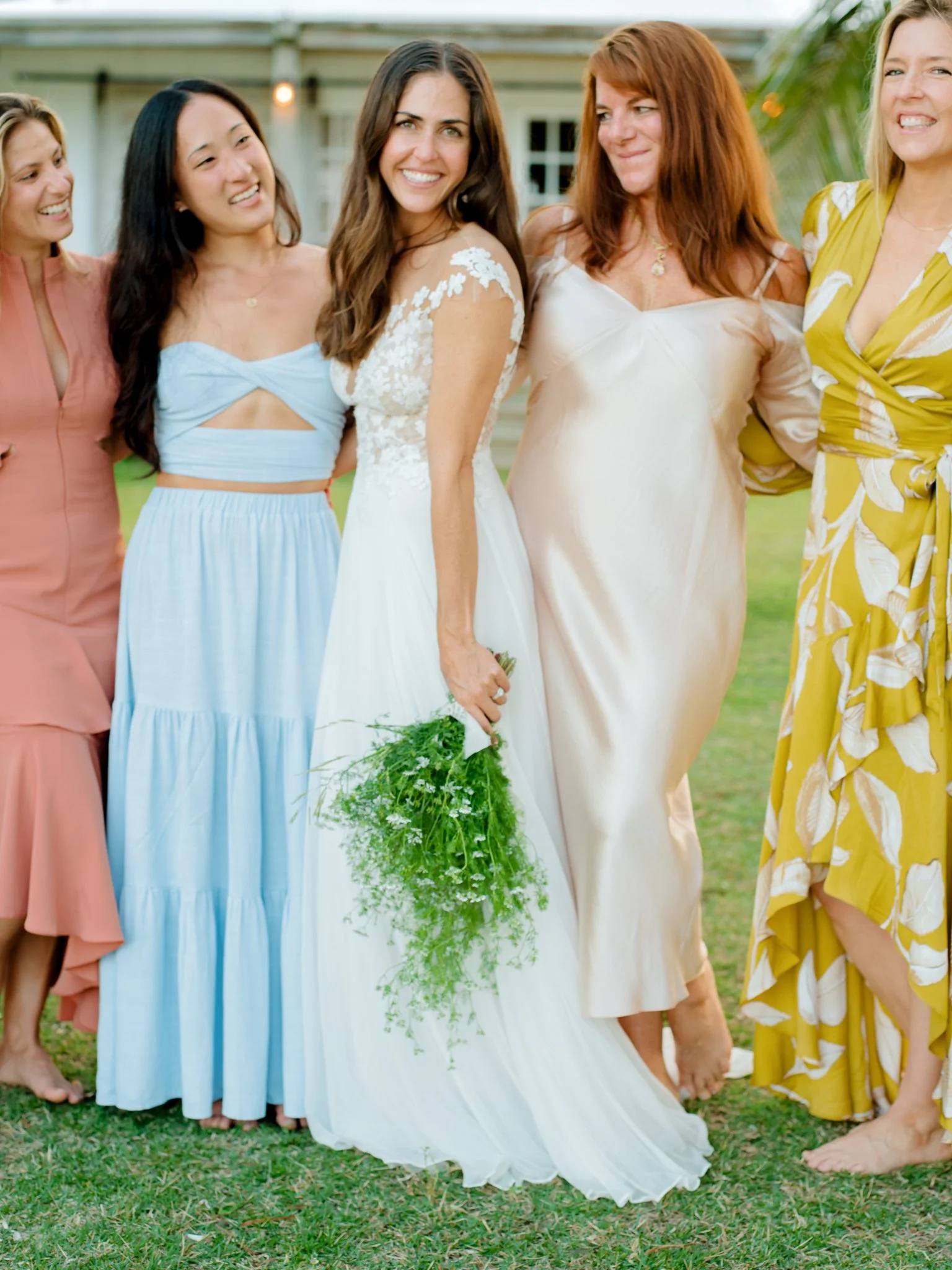 Group of women, including a bride in a white wedding dress holding a green bouquet, standing outdoors on grass.