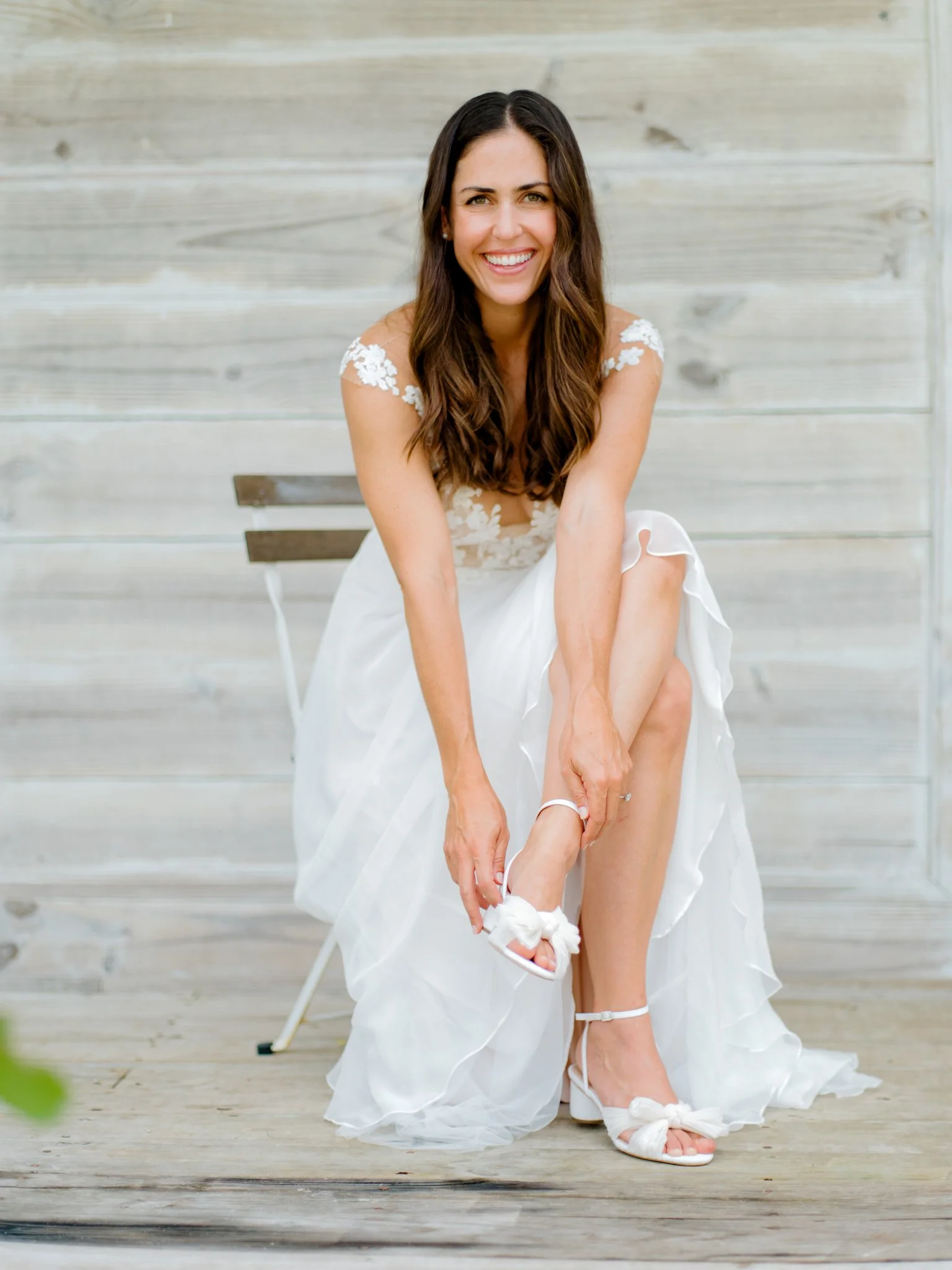 A woman in a wedding dress sitting on a chair, smiling, as she puts on her white high-heeled sandal at The Other Side Hotel Eleuthera during a luxury Bahamas destination wedding.
