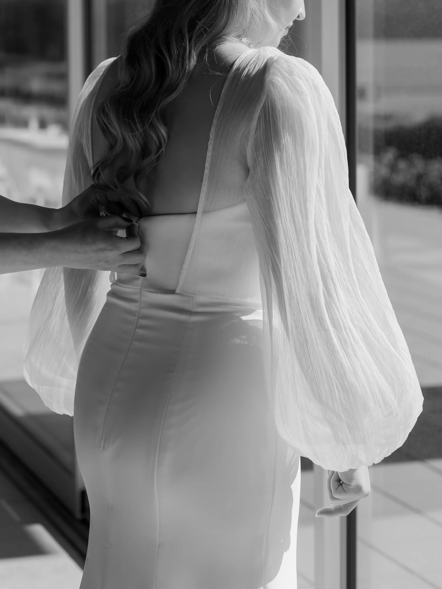 Black and white detail photo of the bride having her gown zipped while standing in natural window light before her Coeur d’Alene wedding.