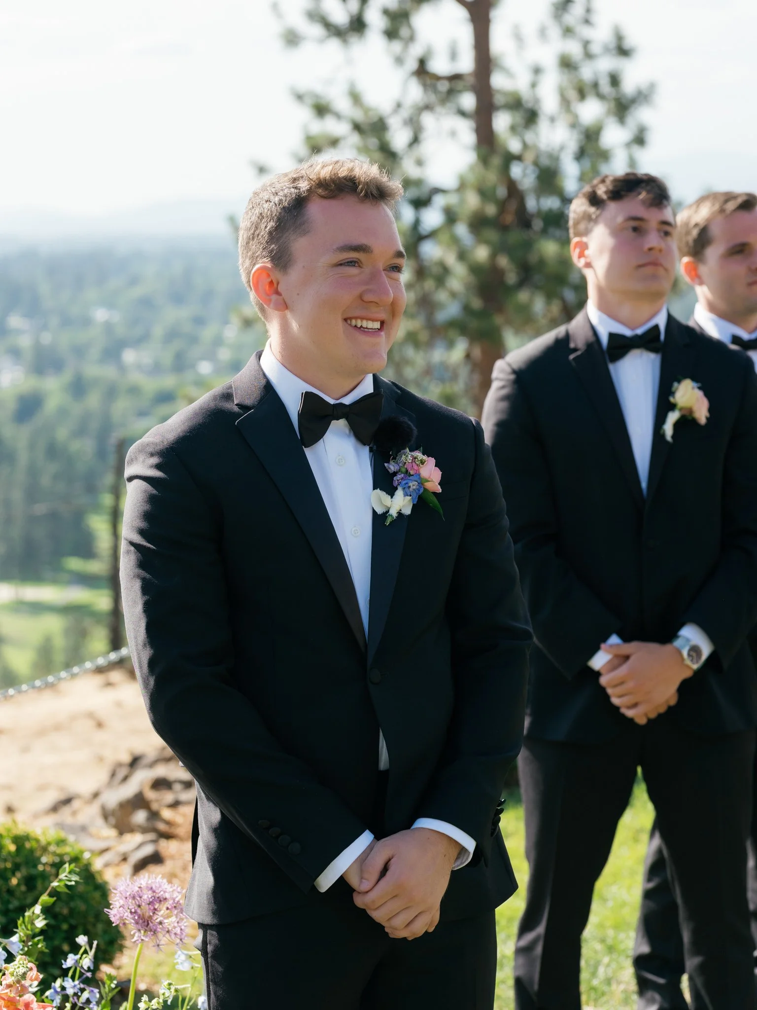 A young man in a black tuxedo with a bow tie, standing outdoors with a slight smile. Other men in tuxedos are visible in the background, and there are trees and a scenic landscape behind them at Elk Point wedding above Lake Coeur d’Alene.
