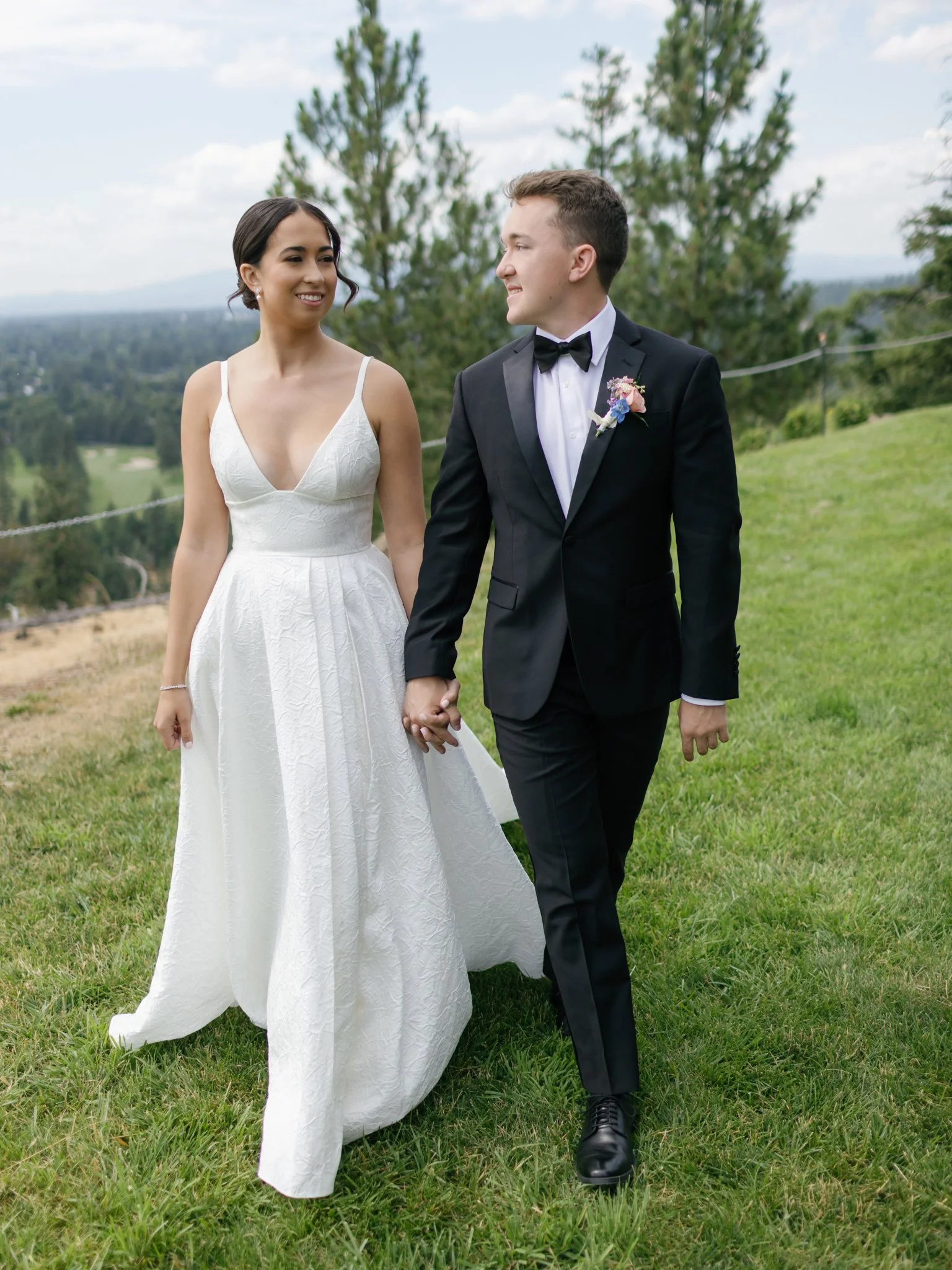 Bride and groom holding hands and walking outdoors on a grassy hill with trees and cloudy sky in the background at Elk Point wedding in Coeur d’Alene.
