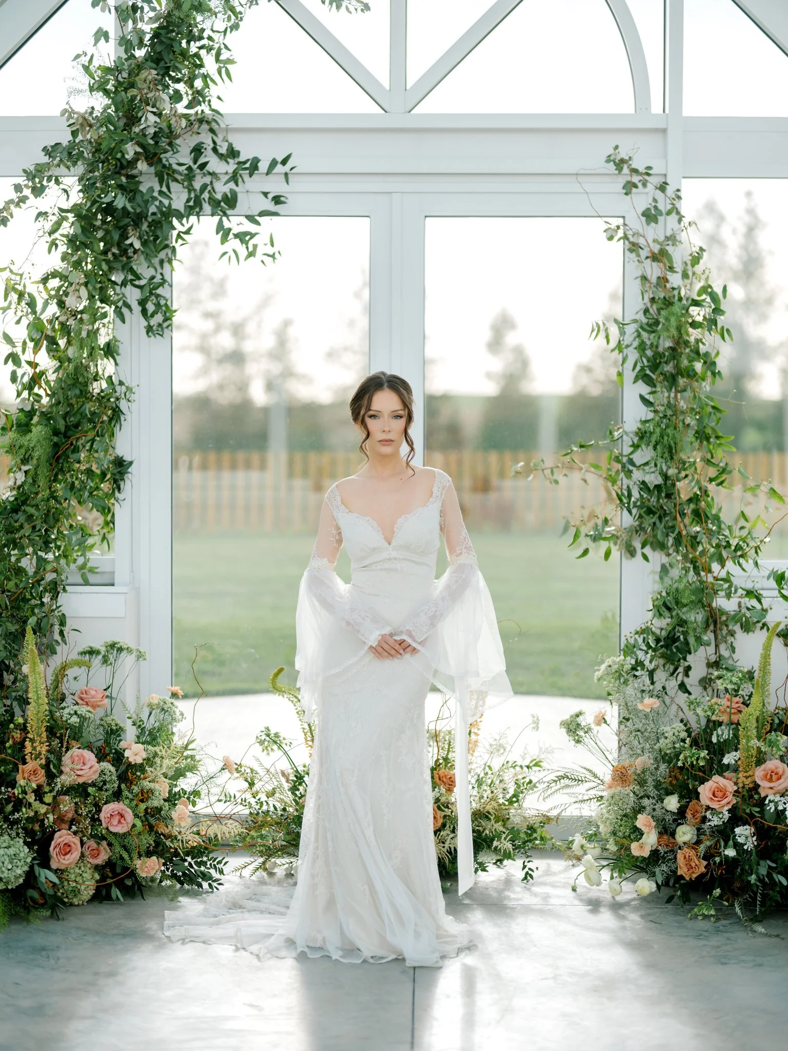 A woman in a white lace wedding dress standing in front of a large glass window with greenery and flowers around her at Greenhouse on Harvard near Spokane, Washington.