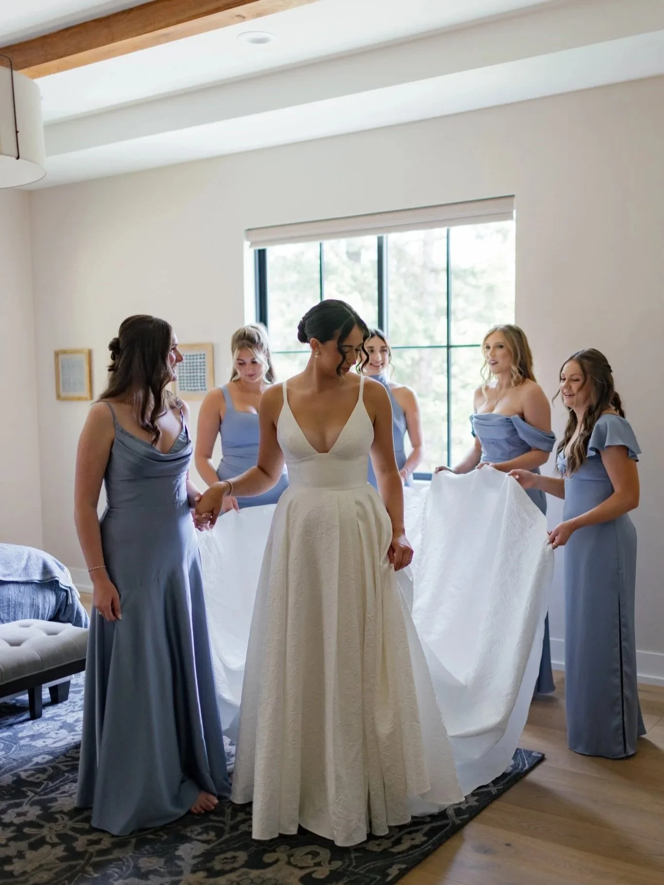 A bride in a white dress surrounded by six women in blue dresses, holding a white cloth, in a bright room with a large window at Elk Point wedding above Lake Coeur d’Alene.
