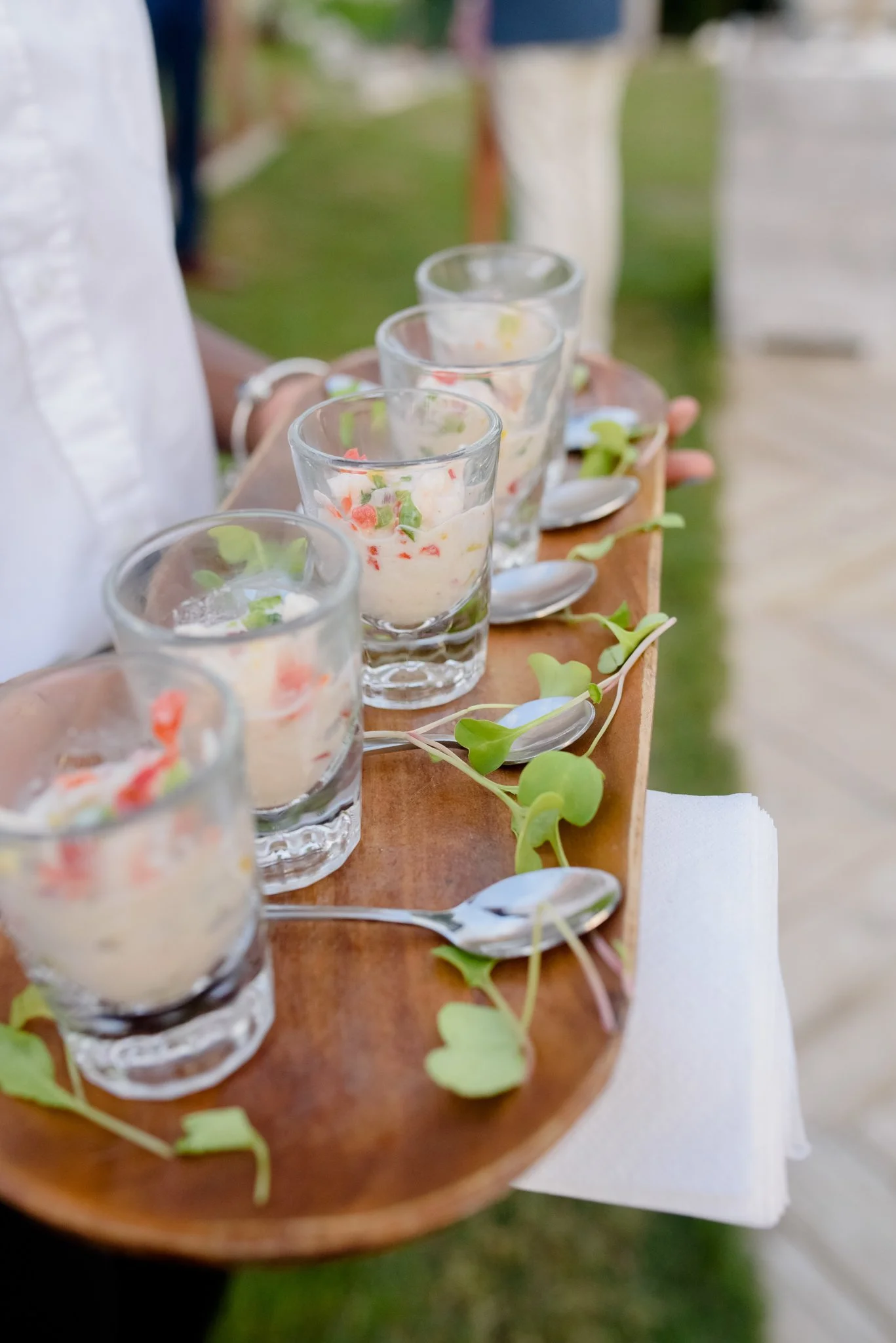 A person holding a wooden tray with four glasses of creamy, garnished dessert and silver spoons, decorated with green leaves, outdoors on a sunny day.