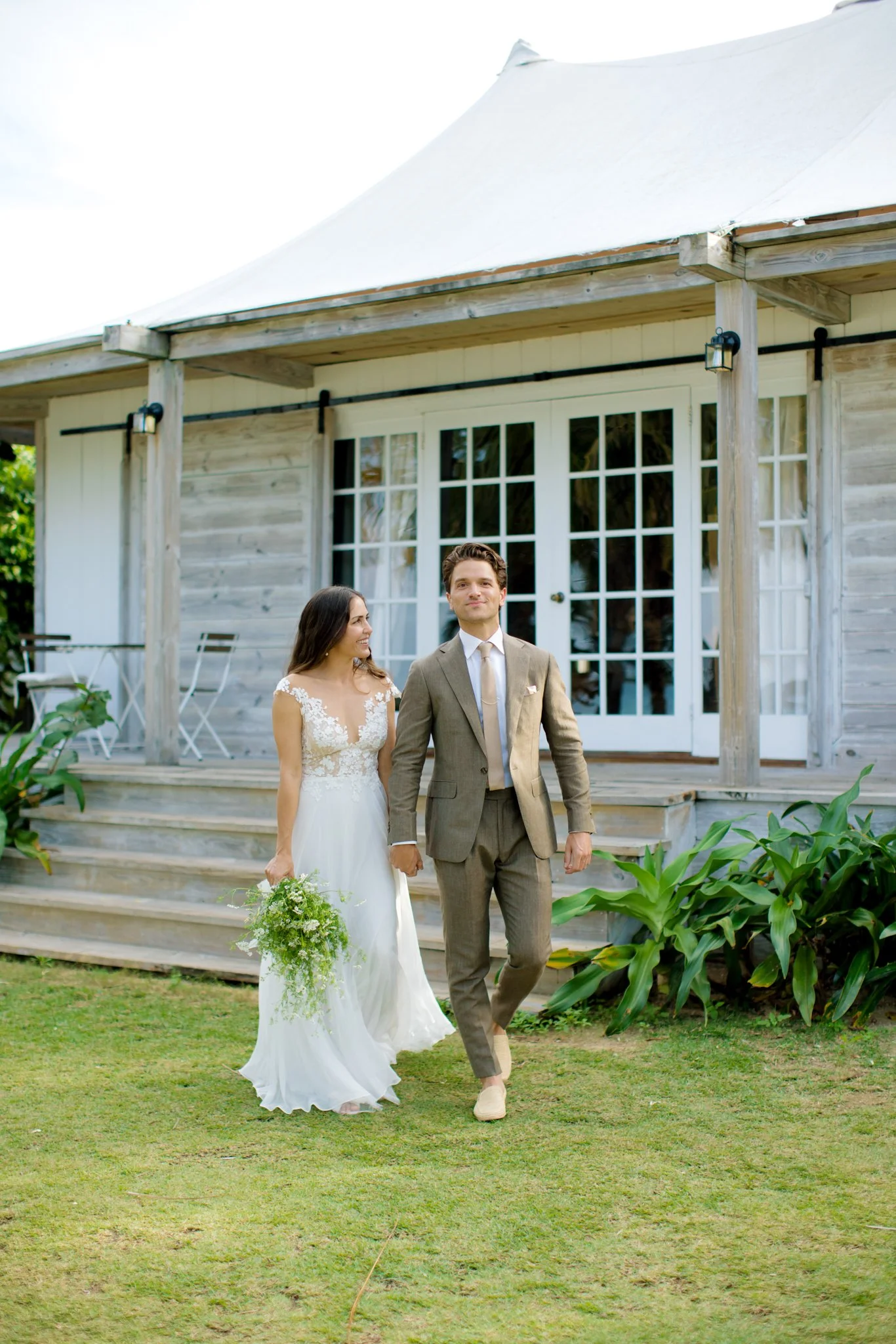 A newlywed couple walking hand in hand outside a wooden house with large glass doors, the bride holding a bouquet of greenery, both smiling, with green plants and grass in the surroundings.