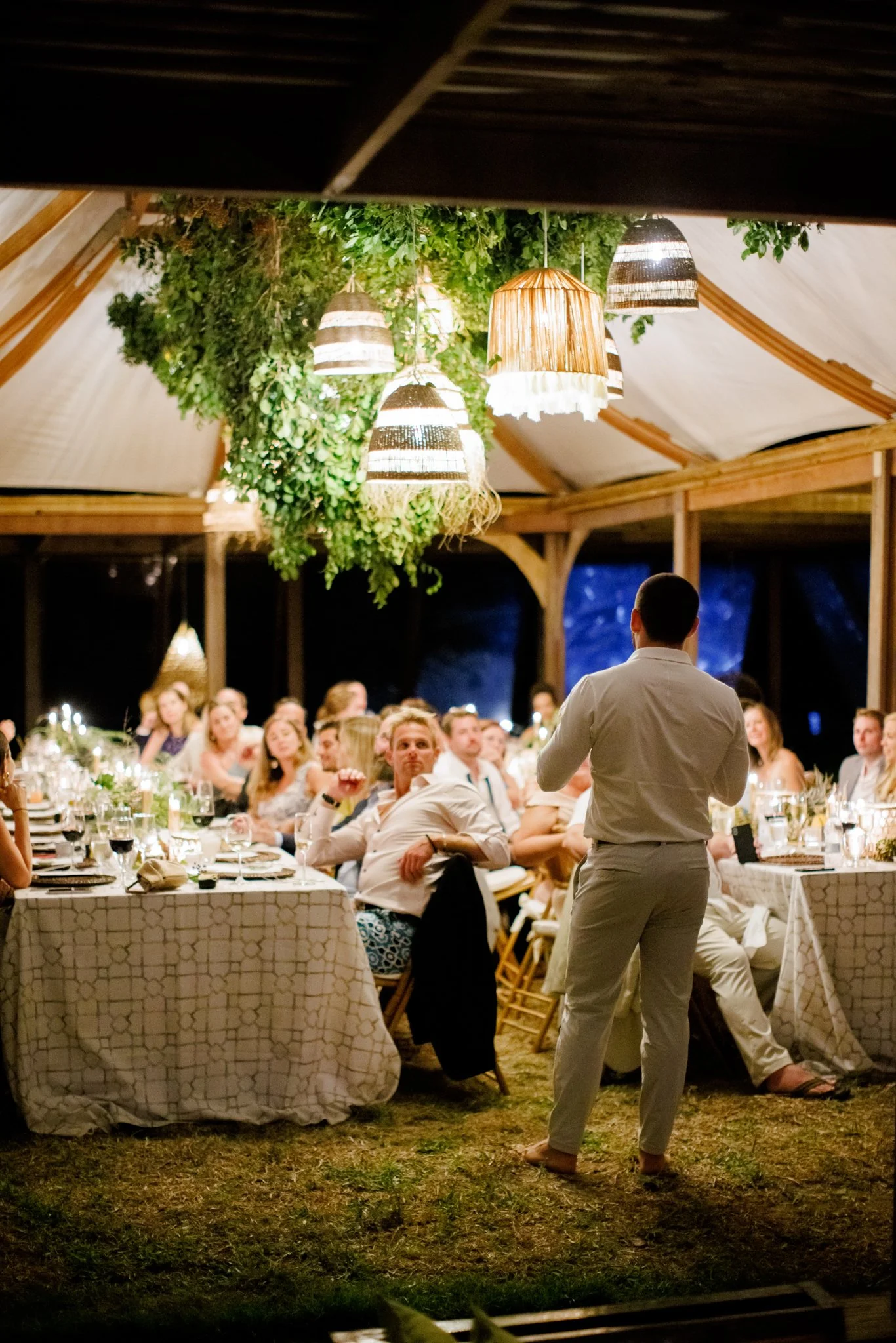 A man giving a speech at a wedding reception under hanging lamps and greenery, with guests seated at long tables watching.