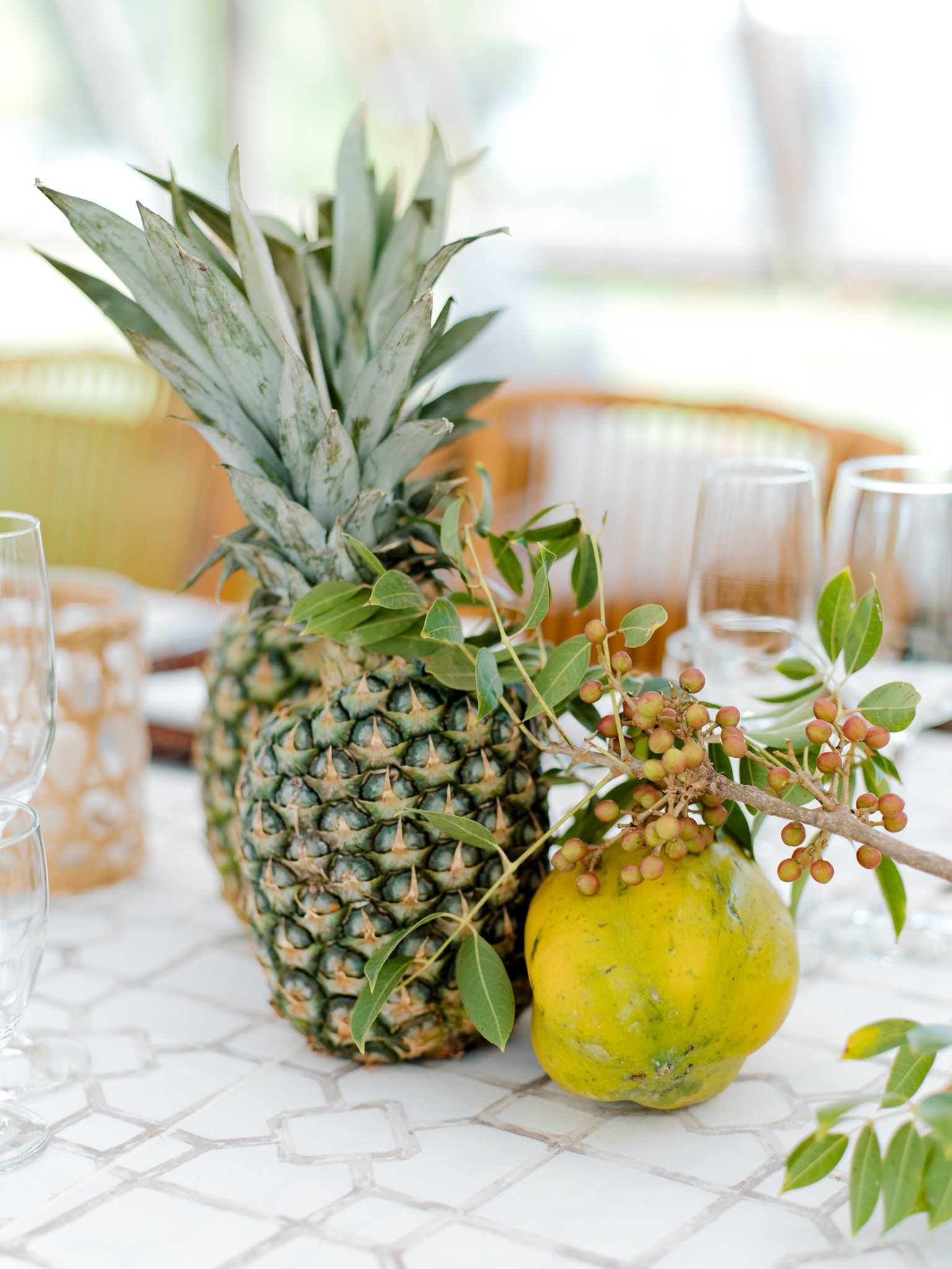 A table centerpiece with a pineapple, a pomegrante, and assorted greenery on a white tablecloth at The Other Side Hotel Eleuthera during a luxury Bahamas destination wedding.