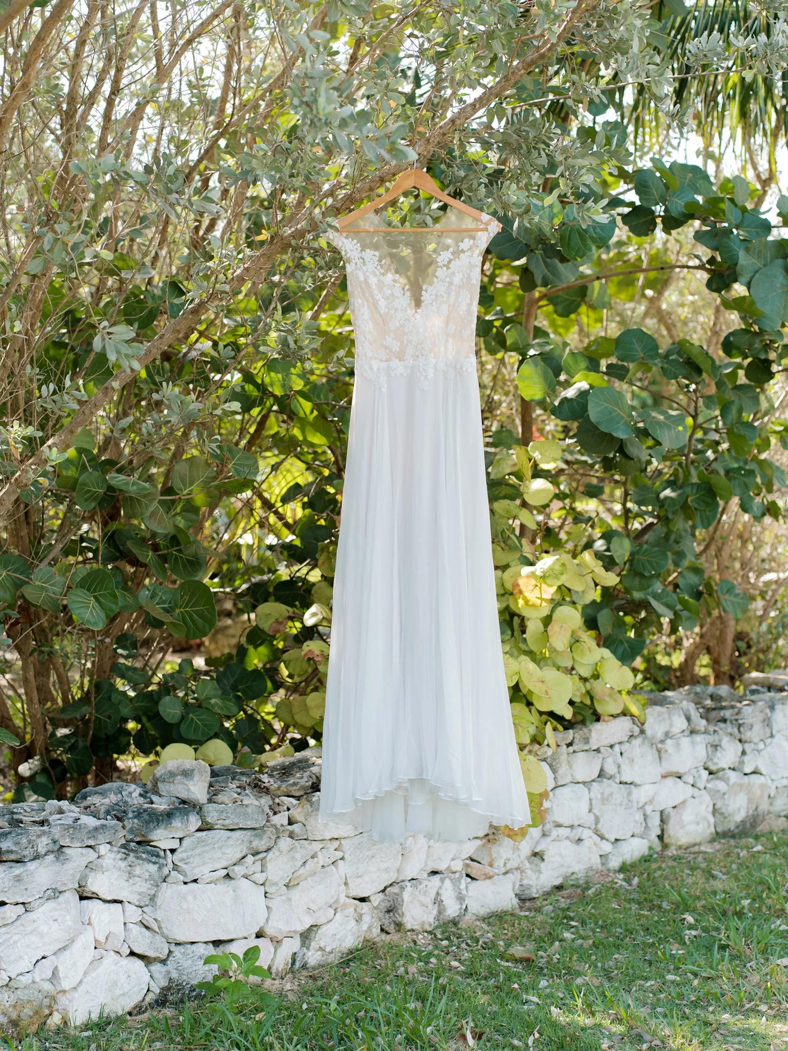 A white wedding dress with lace bodice hanging from a hanger on a tree branch outdoors, with green foliage and a stone border in the background.