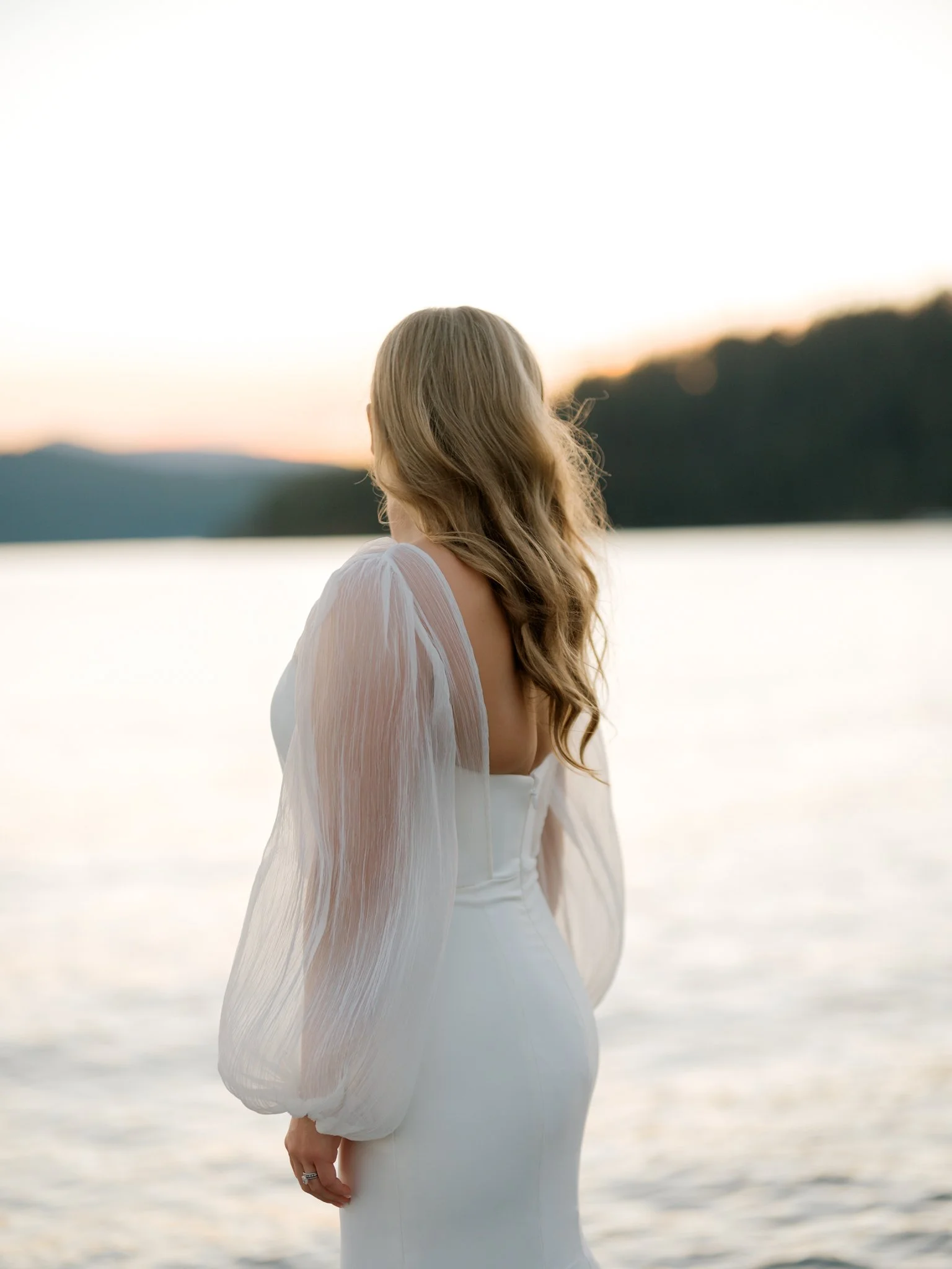 A woman in a white dress with sheer, puffy sleeves standing by a body of water at sunset, with her back facing the camera and her long, wavy hair flowing down at the Hagadone Event Center in Coeur d'Alene, Idaho.