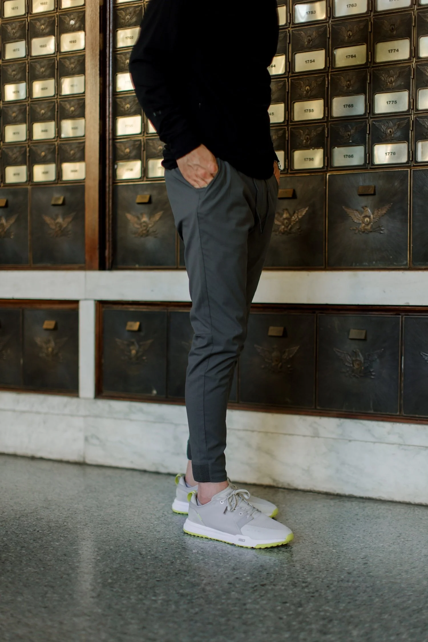A person dressed in a black jacket, gray pants, and white sneakers standing in front of a wall of postal mailboxes.