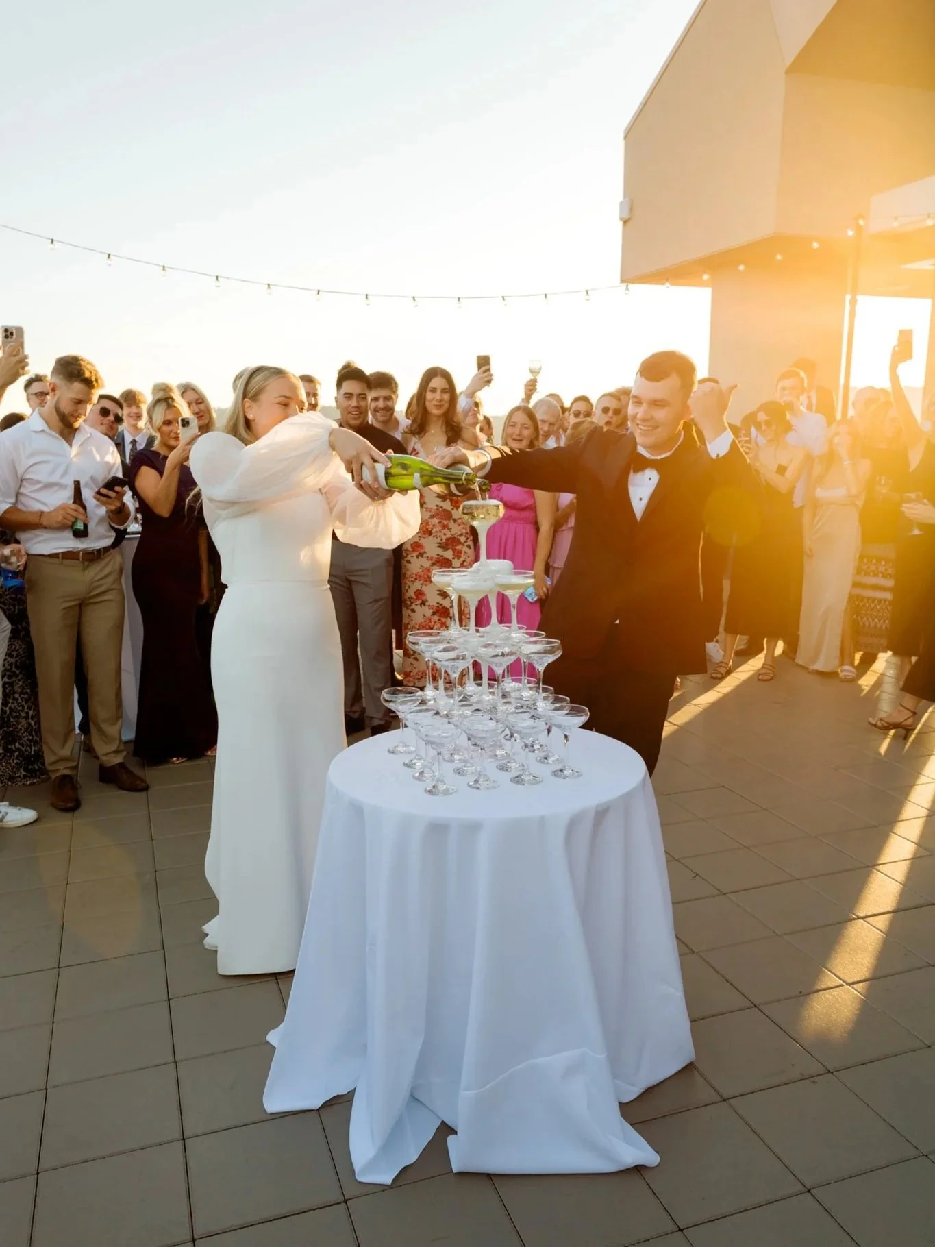 A newlywed couple pouring champagne into a pyramid of glasses at their wedding reception, with guests gathered around watching and celebrating during sunset.