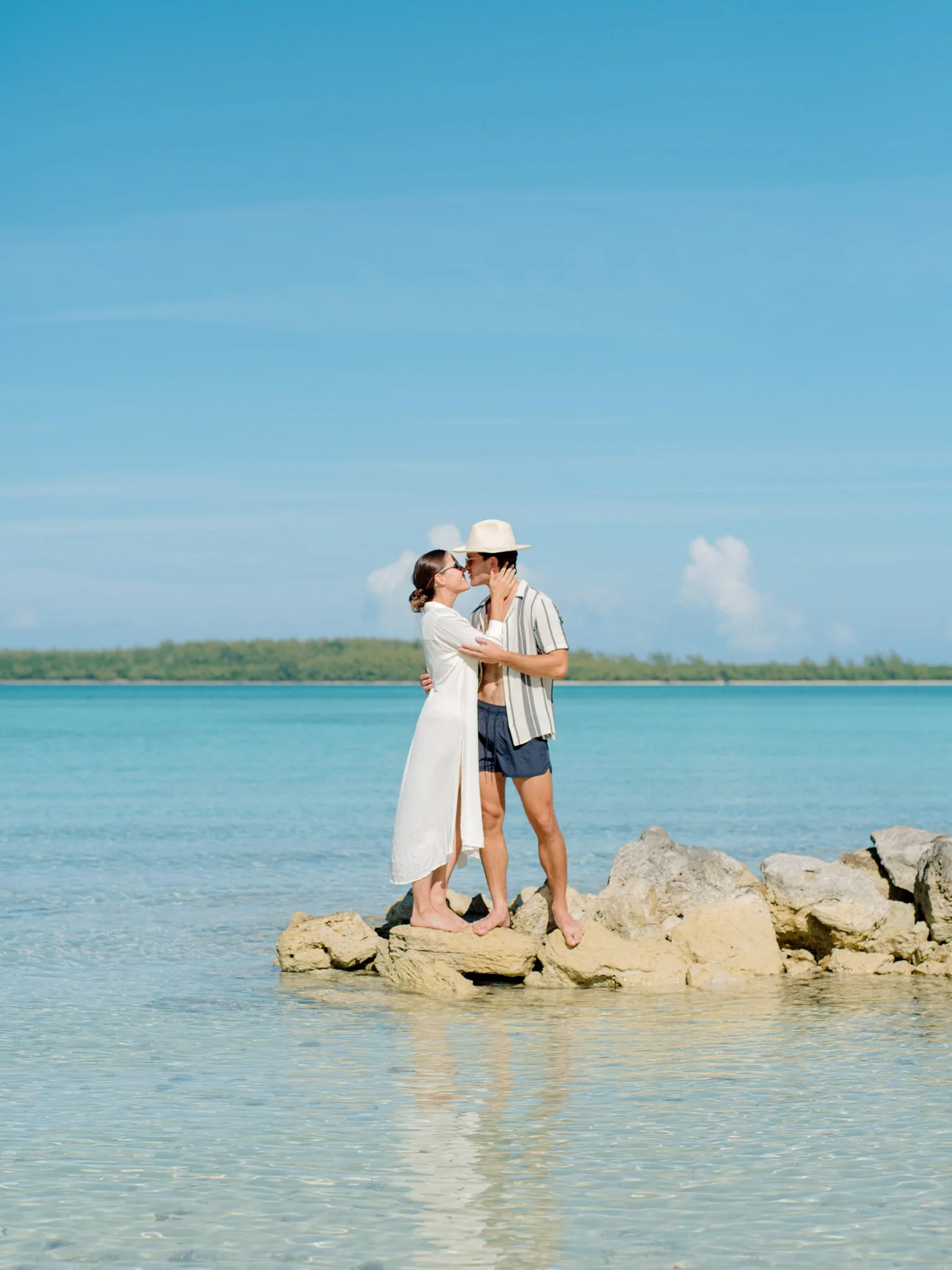 A couple standing on rocks at the beach, sharing a kiss with the ocean and a clear blue sky in the background at The Other Side Hotel during their destination wedding in Eleuthera, Bahamas.