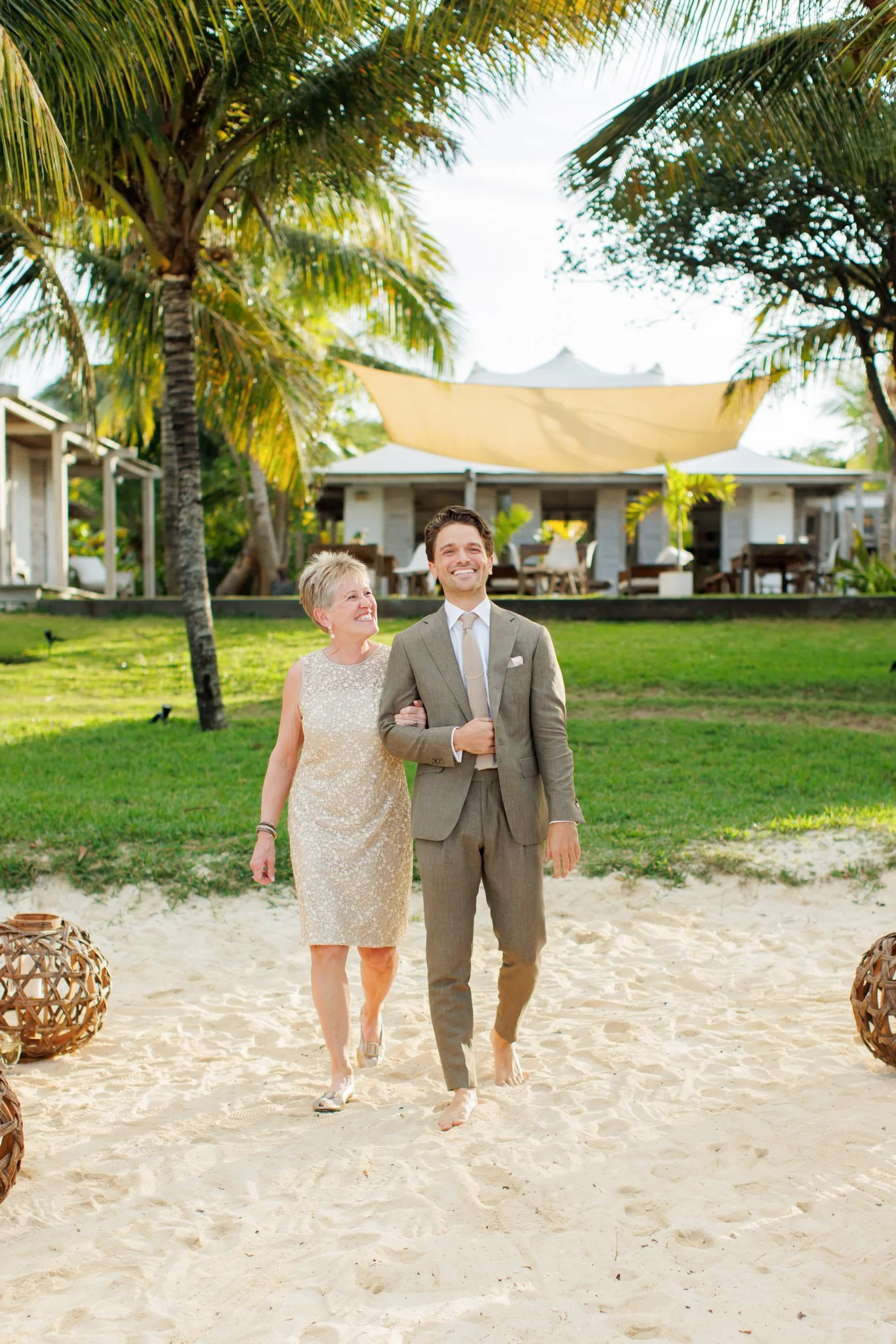 A couple, an older woman and a young man, walking barefoot on a sandy beach, smiling, with tropical trees and a large house or resort in the background at The Other Side Hotel Eleuthera during a luxury Bahamas destination wedding.