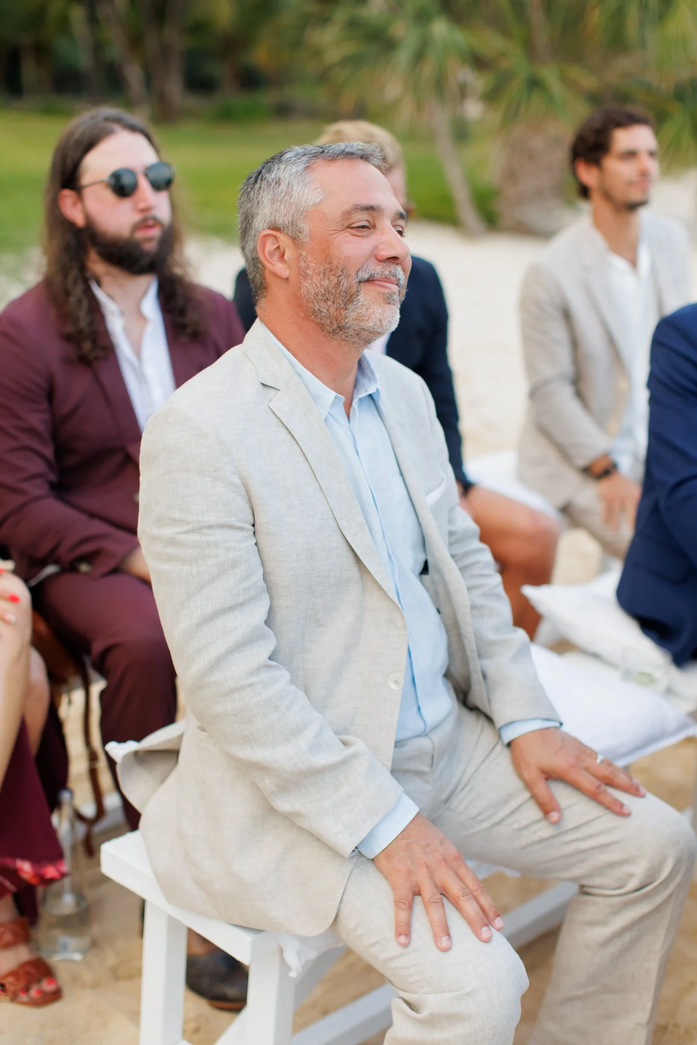 A man with gray hair and a beard is sitting outdoors in a light-colored suit during a formal event, with other people in the background also dressed in formal attire.