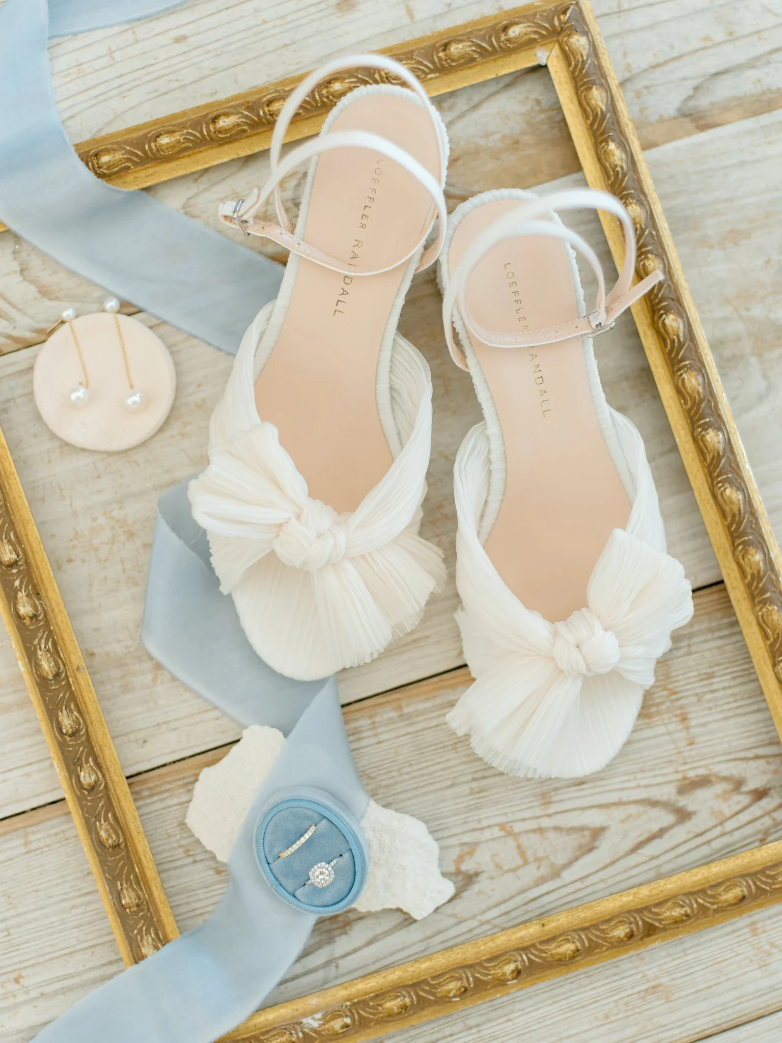 A pair of white pleated ruched-strap high-heeled shoes with knotted bows on the toes, placed in a gold ornate picture frame on a wooden surface. An ivory circular earrings display with pearl-stud earrings and a tied blue fabric ribbon with a ring inside are also visible.
