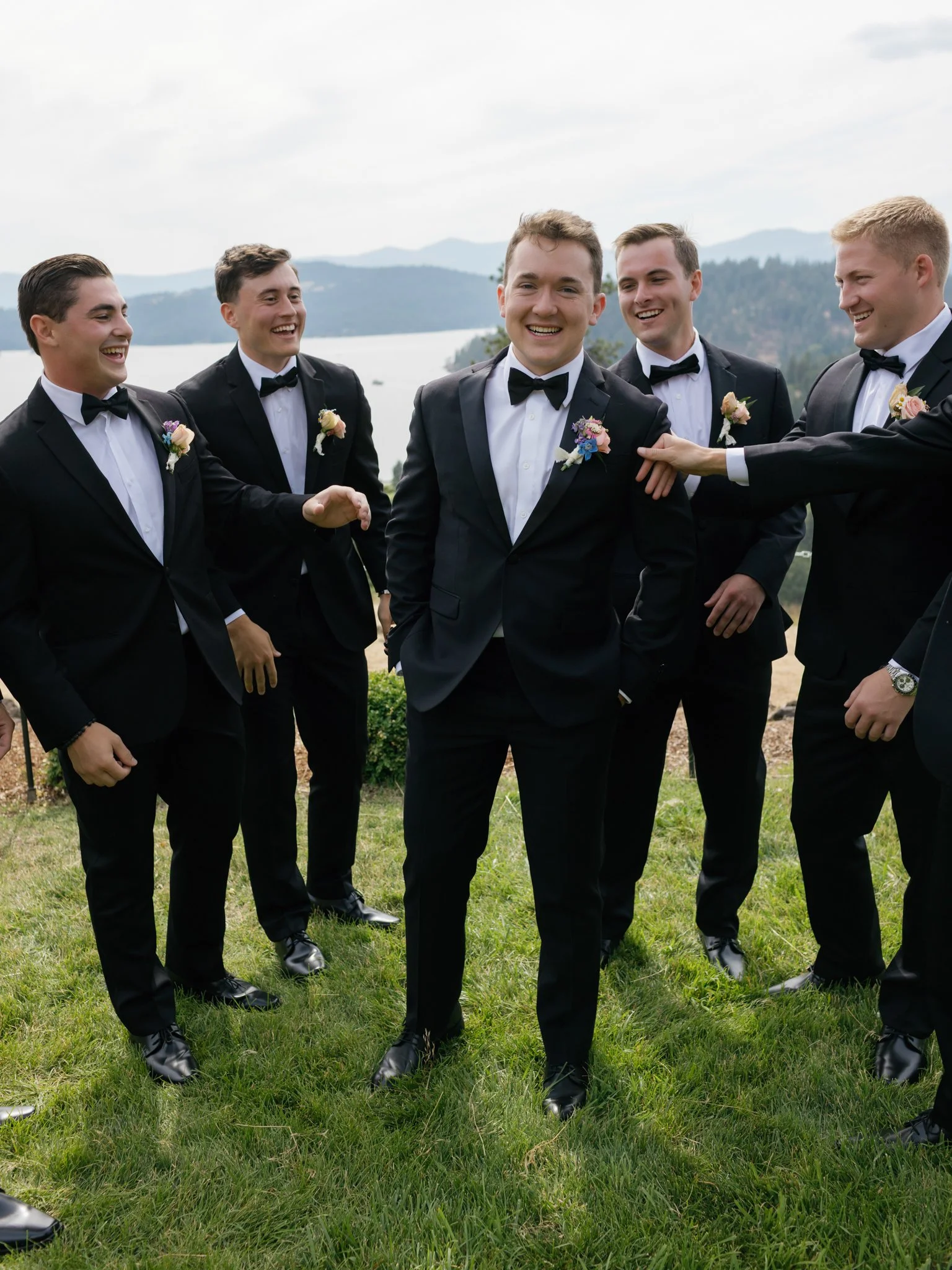 Group of men in tuxedos standing on grass outdoors, smiling and laughing, with a lake and mountains in the background at Elk Point wedding above Lake Coeur d’Alene.
