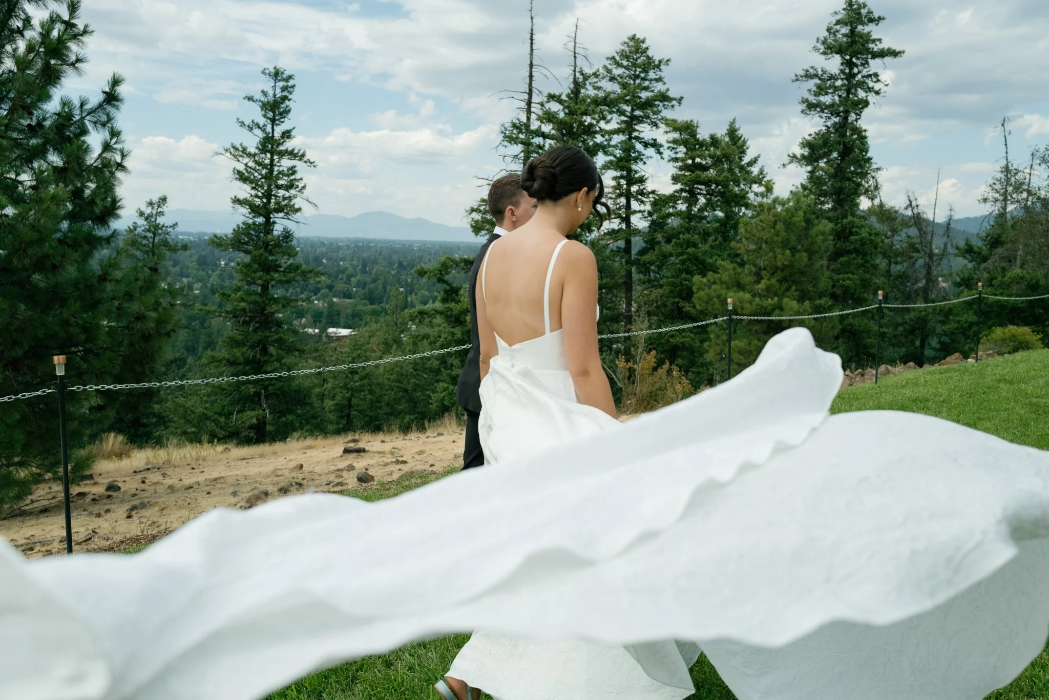 A bride and groom walking outdoors on a grassy hill with trees and mountains in the background during a wedding celebration at Elk Point wedding in Coeur d’Alene.
