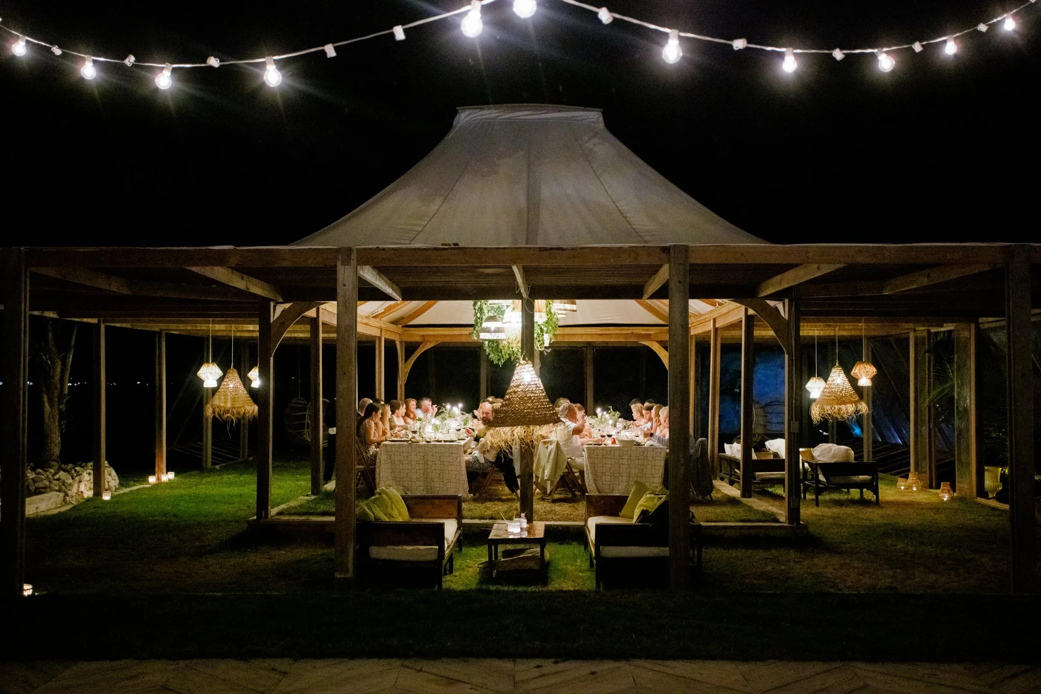 Nighttime outdoor dinner party under a large central tent with string lights, surrounded by a wooden gazebo with hanging lamps and candles, filled with people seated at tables.
