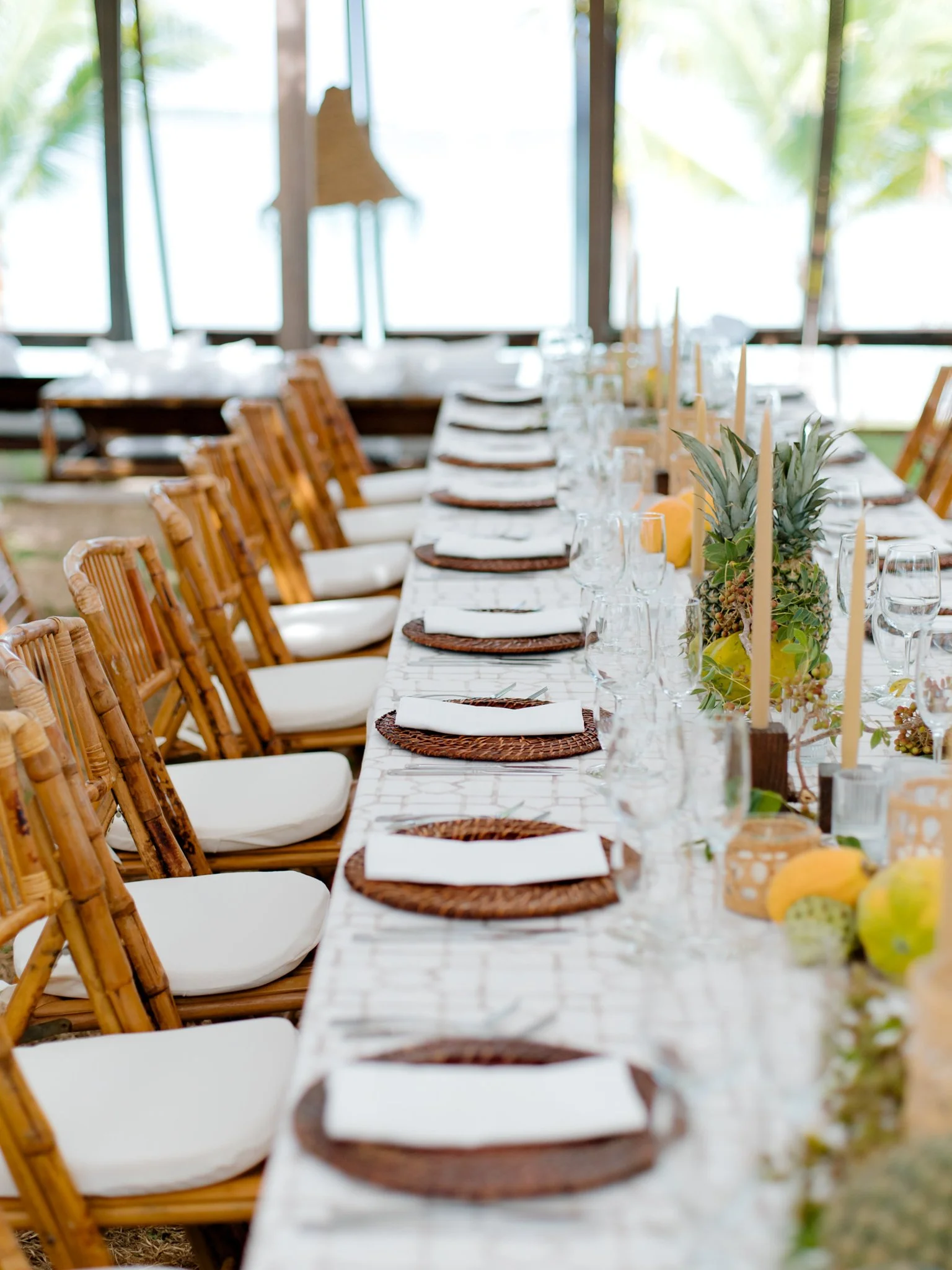Long dining table set for a tropical celebration, with white napkins on dark wicker placemats, surrounded by wooden chairs with white cushions, and decorated with fruit, candles, and a floral centerpiece, near large windows showing palm trees outside.